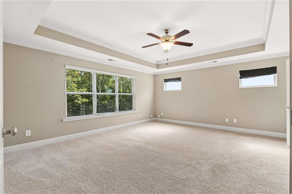 Empty carpeted bedroom with a tray ceiling, ceiling fan, and wide windows.