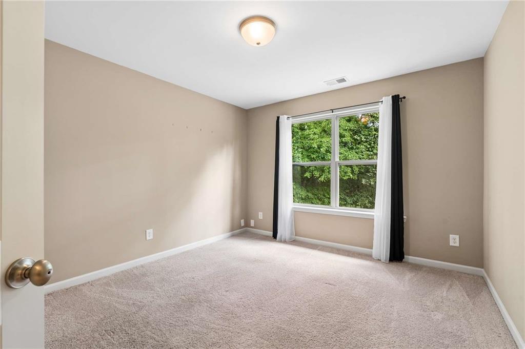 Empty bedroom with beige walls, carpet, and a curtained window overlooking trees.