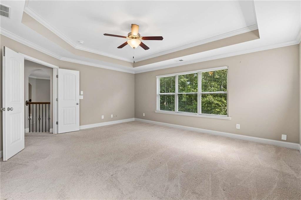 Empty carpeted bedroom with a tray ceiling, ceiling fan, and wide window overlooking greenery.