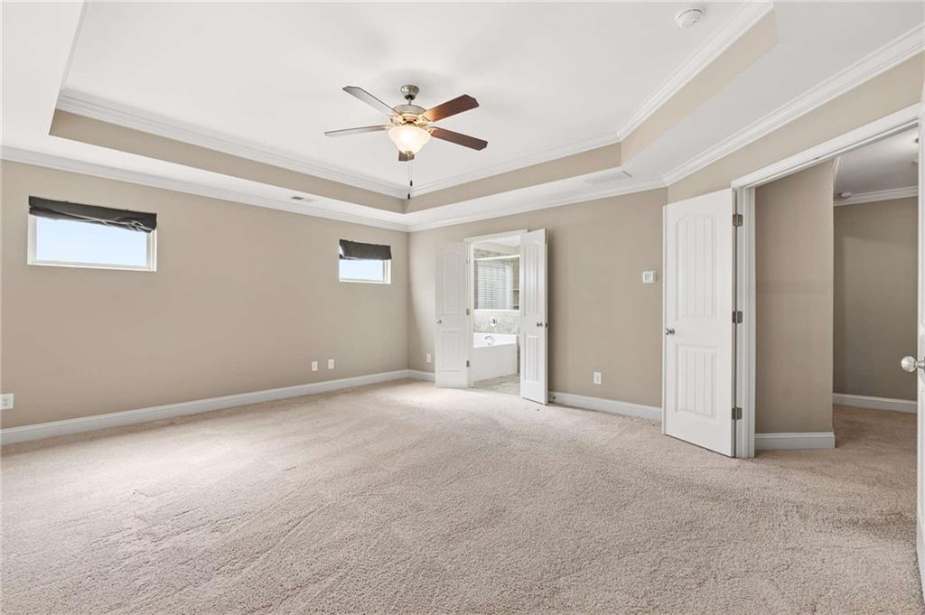 Empty bedroom with beige carpet, a tray ceiling, ceiling fan, and doorway to an adjoining bathroom.