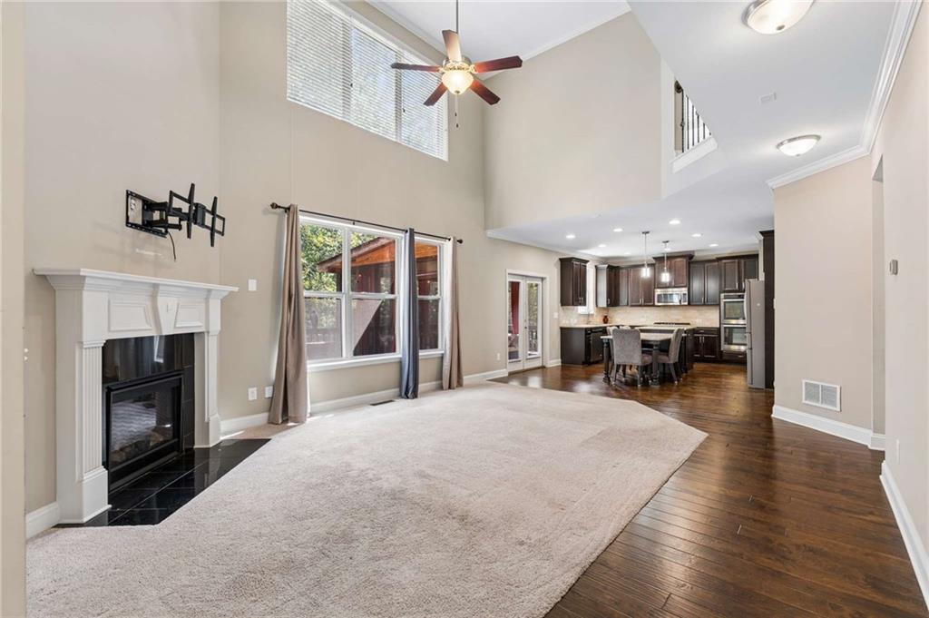 Two-story living room with a white fireplace, tall windows, a ceiling fan, and an open kitchen.