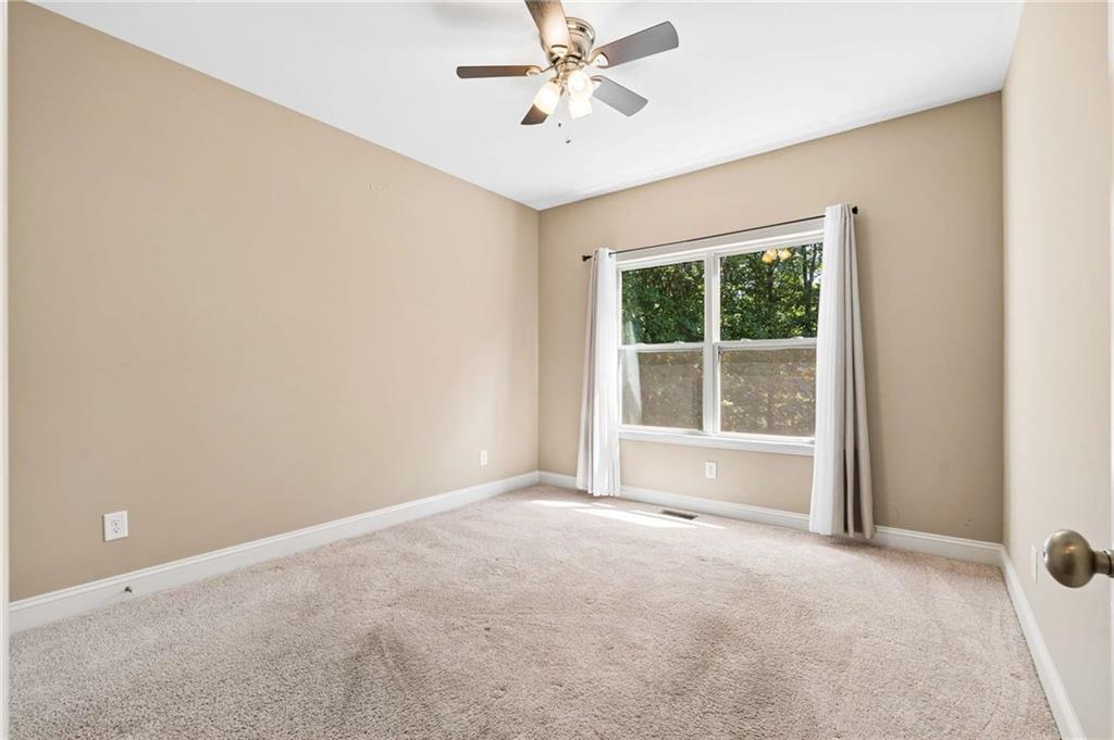 Empty bedroom with beige walls, light carpet, and a curtained window overlooking trees.