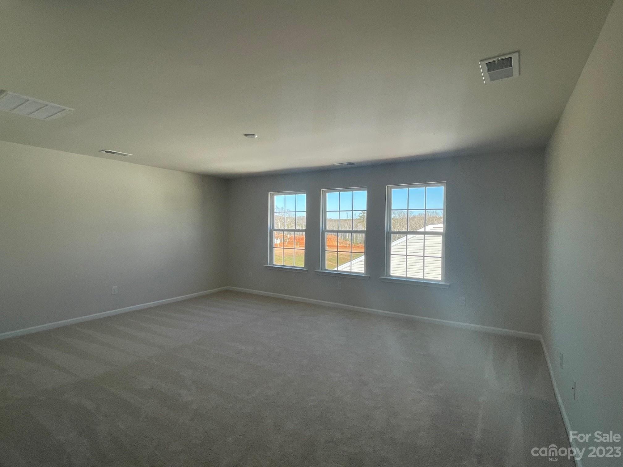 Empty carpeted room with three tall windows and recessed ceiling lights.