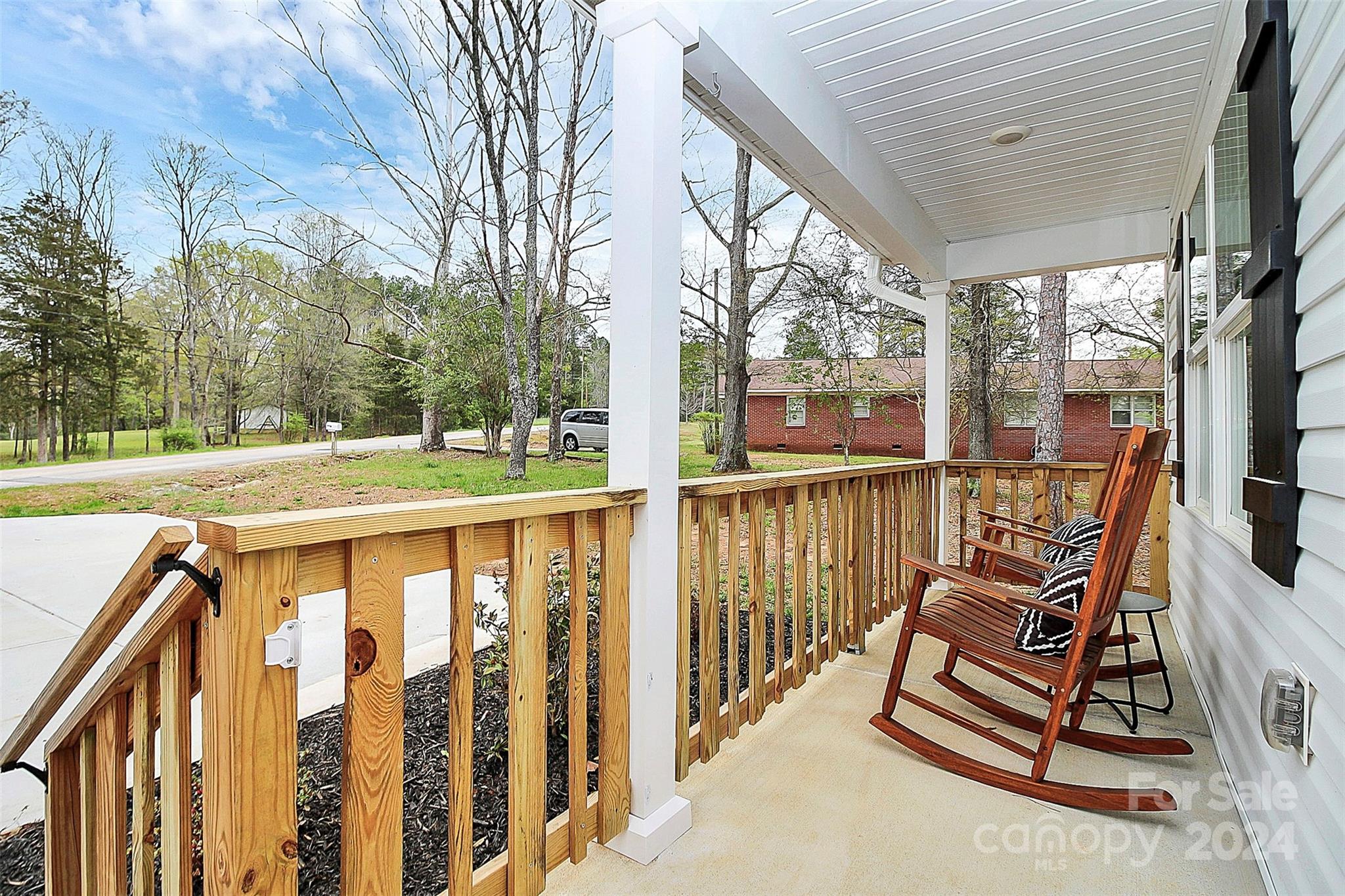 Covered front porch with wooden railing and two rocking chairs overlooking a grassy yard and road.