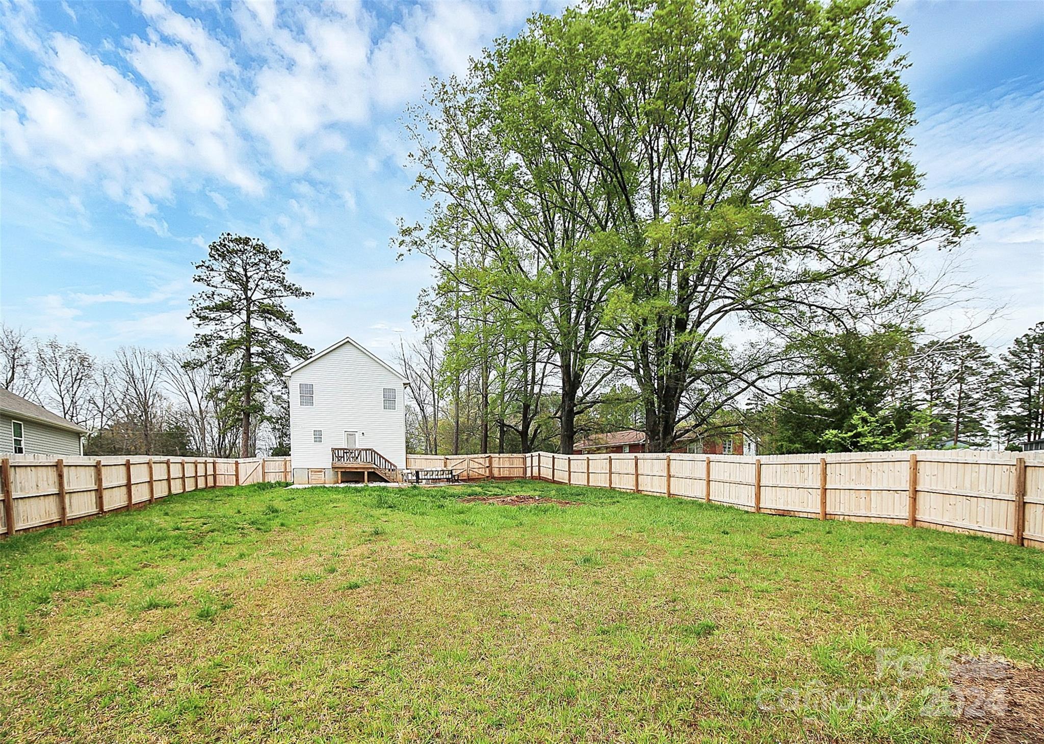 Home’s fenced backyard with a broad lawn, small deck, and mature trees.
