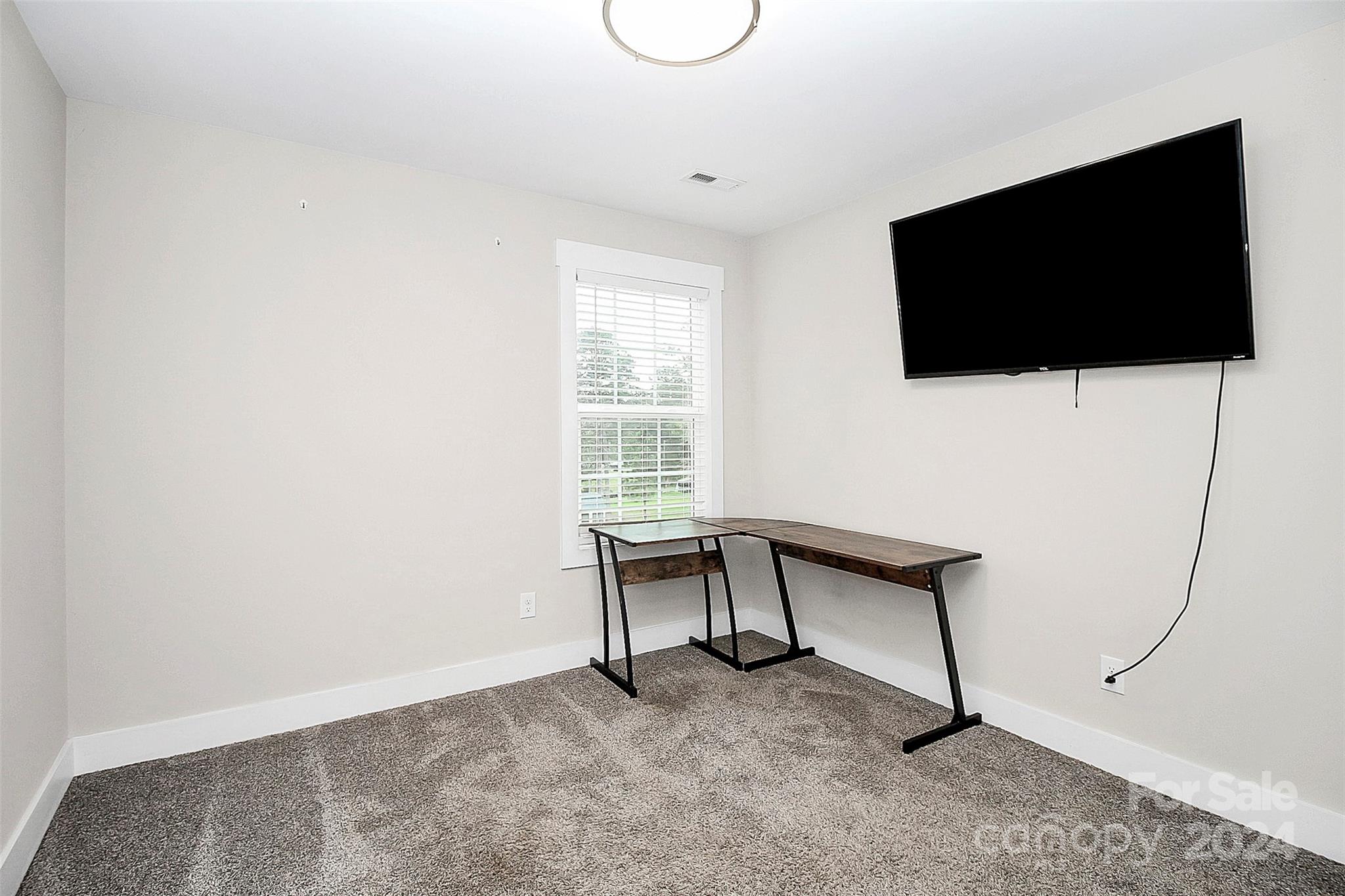 Carpeted home office with a corner desk, wall-mounted television, and window with white blinds.