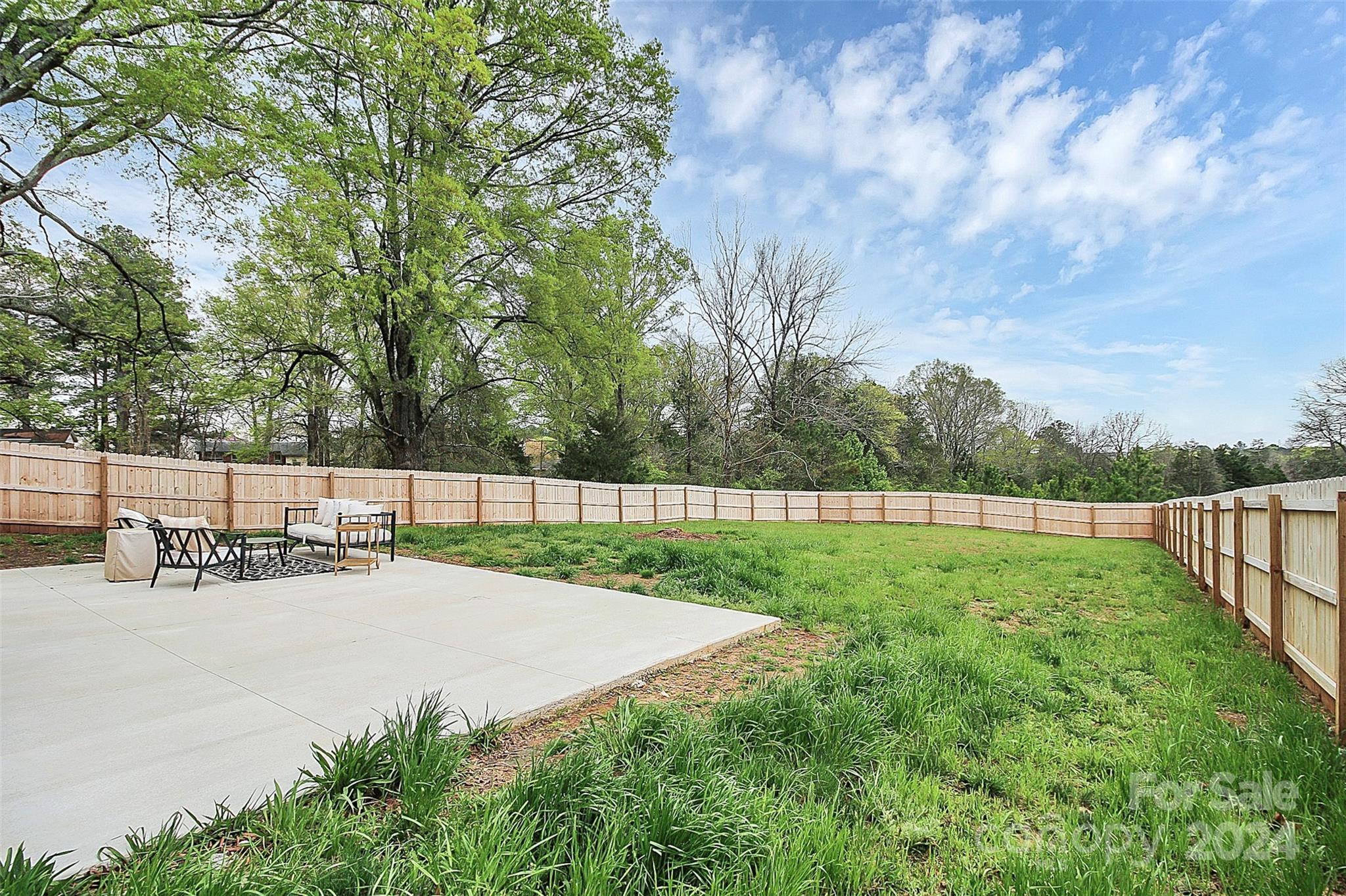 Fenced backyard with a concrete patio, outdoor seating, and a broad green lawn.