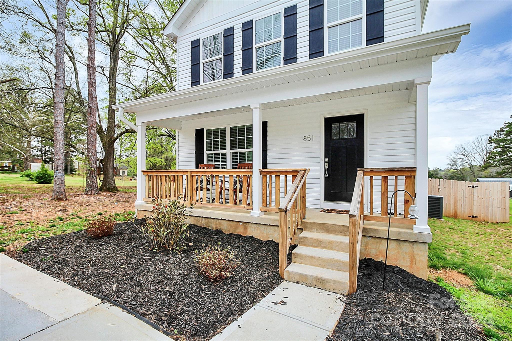 White two-story house with a covered wooden-railed porch, black shutters, and landscaped front yard.