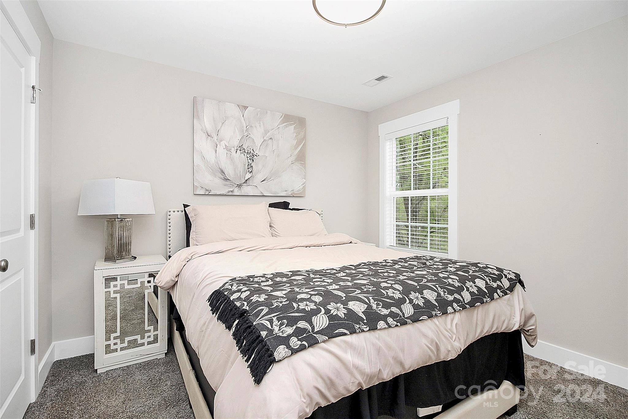 Furnished bedroom with pale bedding, a dark floral throw, and a window with white blinds.