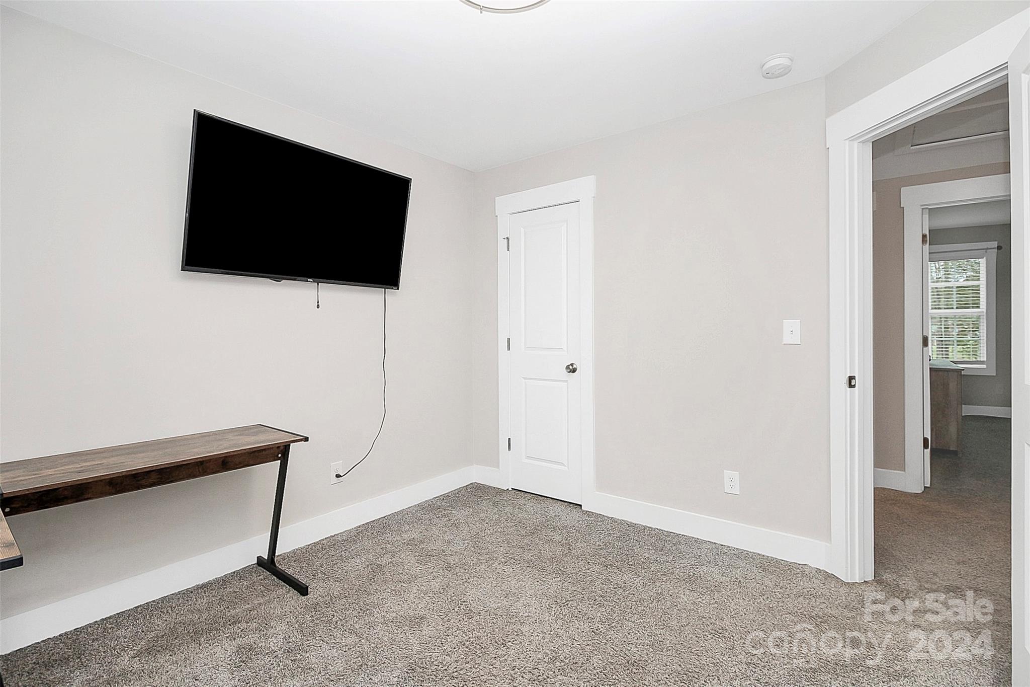 Carpeted room with a wall-mounted television, narrow desk, and doorway to an adjoining room.