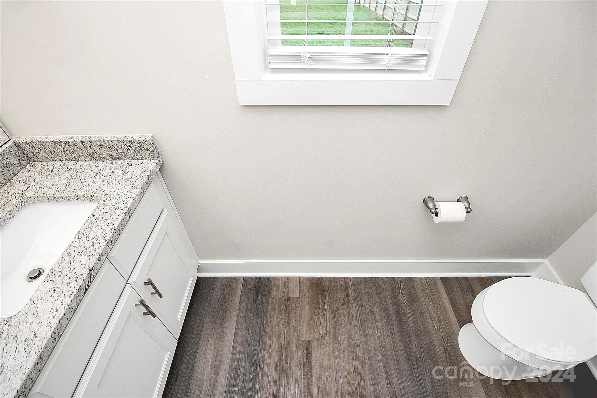 Bathroom with a granite vanity, toilet, window, and dark wood-look flooring.