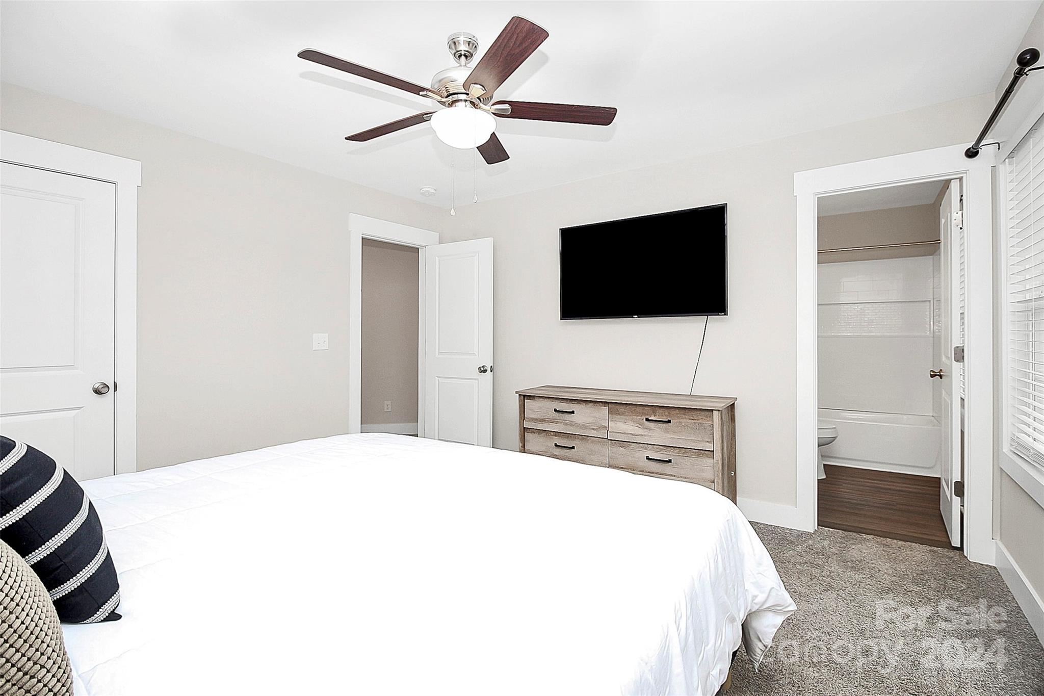 Furnished bedroom with a white bed, ceiling fan, and open doorway to an attached bathroom.