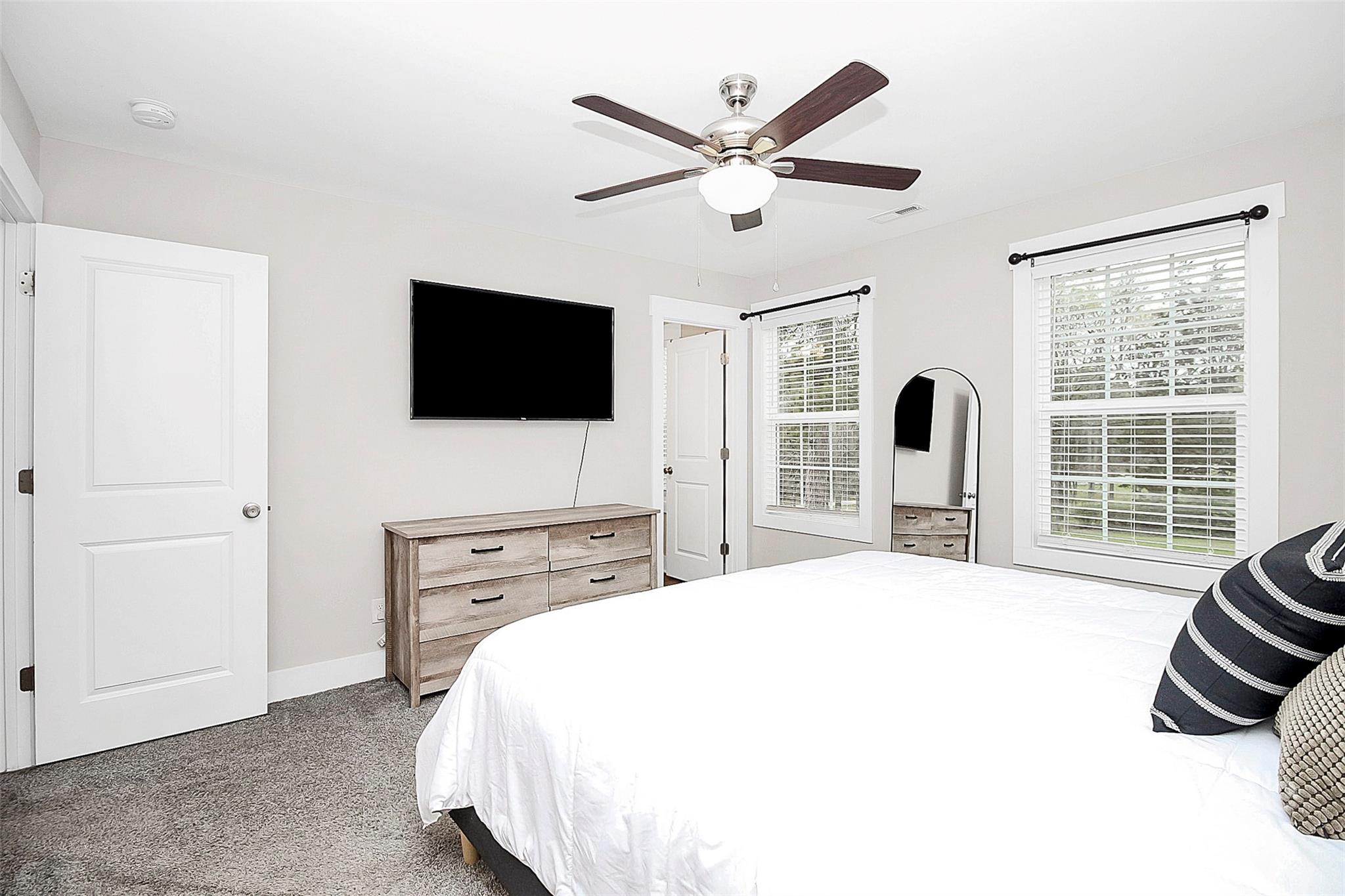 Bedroom with a white bed, gray carpet, ceiling fan, and wall-mounted television.