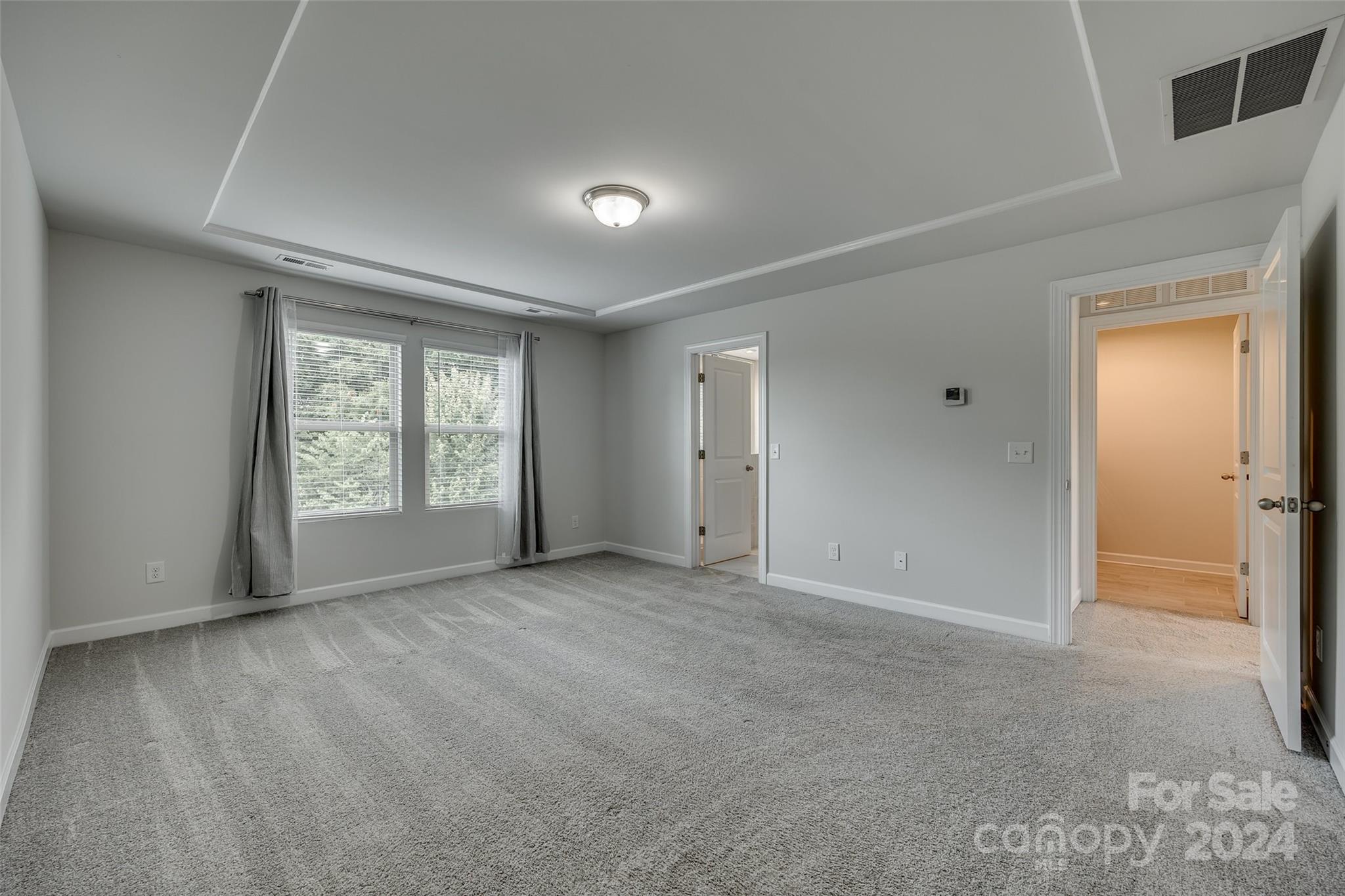 Empty carpeted bedroom with a tray ceiling, two curtained windows, and open doorways.