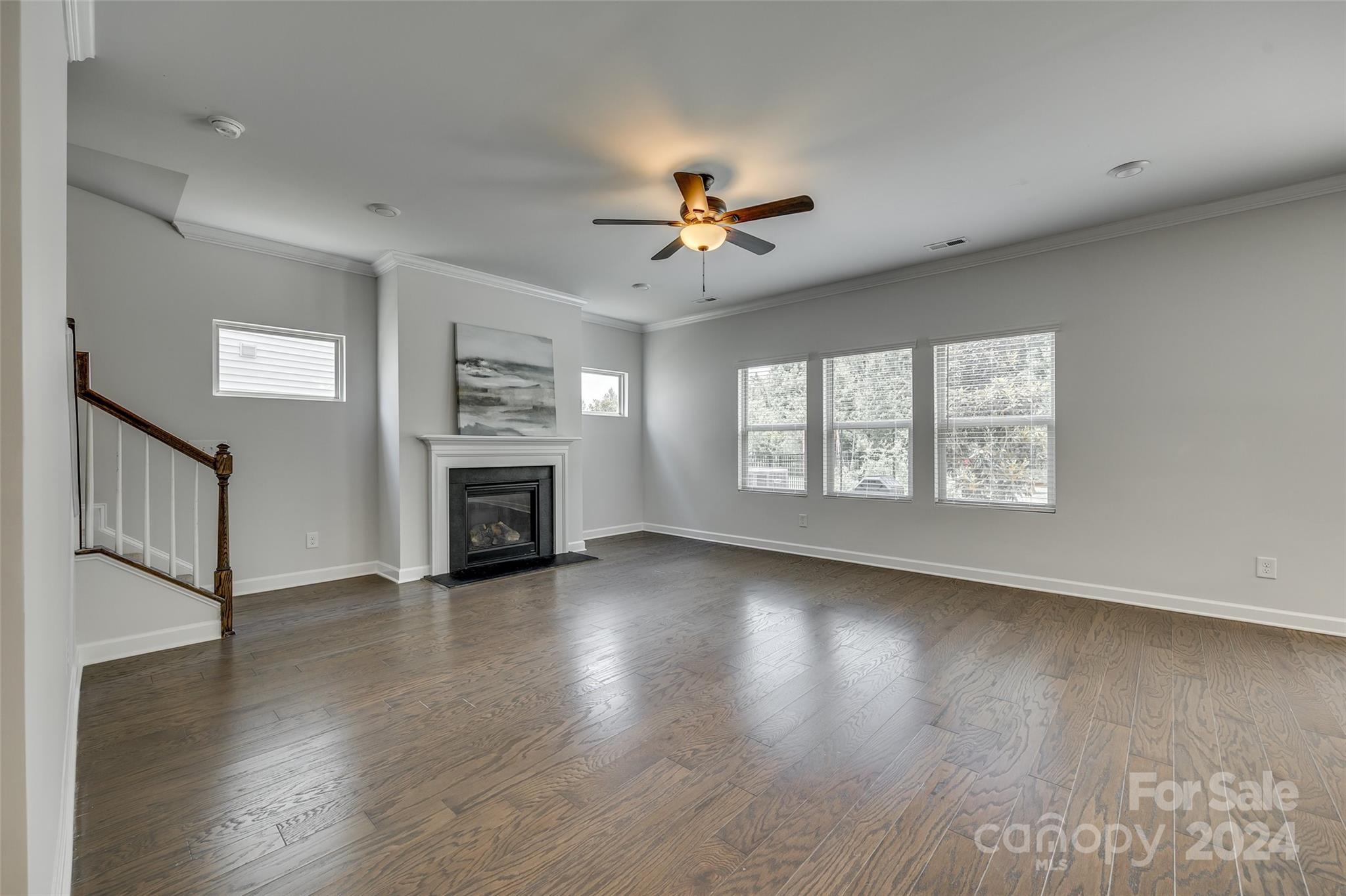 Empty living room with hardwood floors, a fireplace, ceiling fan, and staircase.