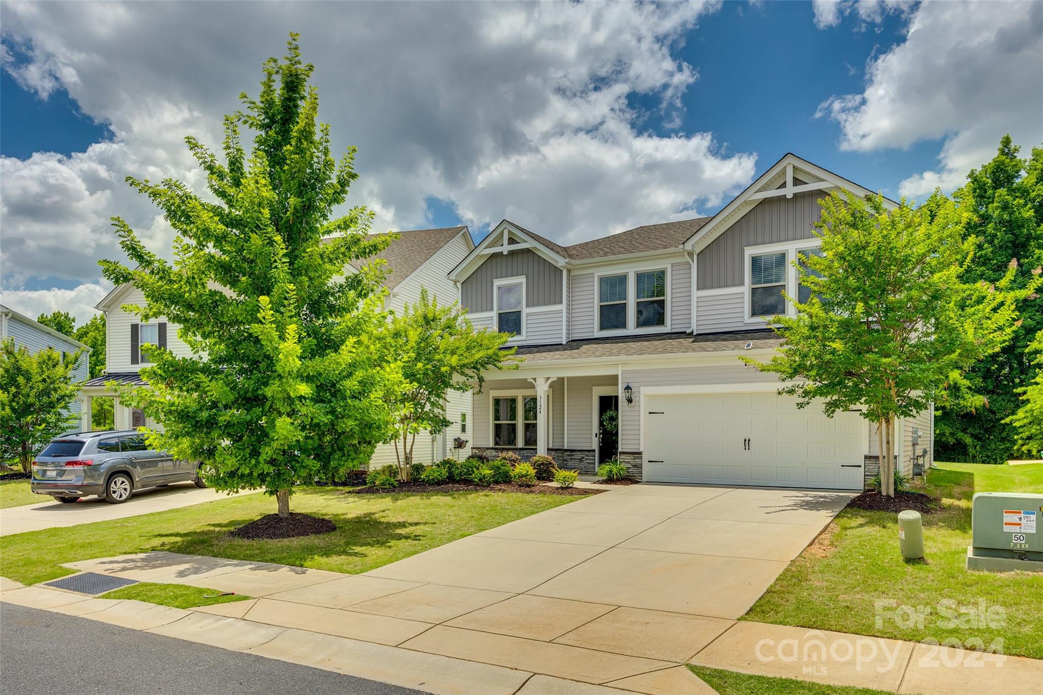 Two-story gray and white house with a two-car garage, wide driveway, and mature trees.