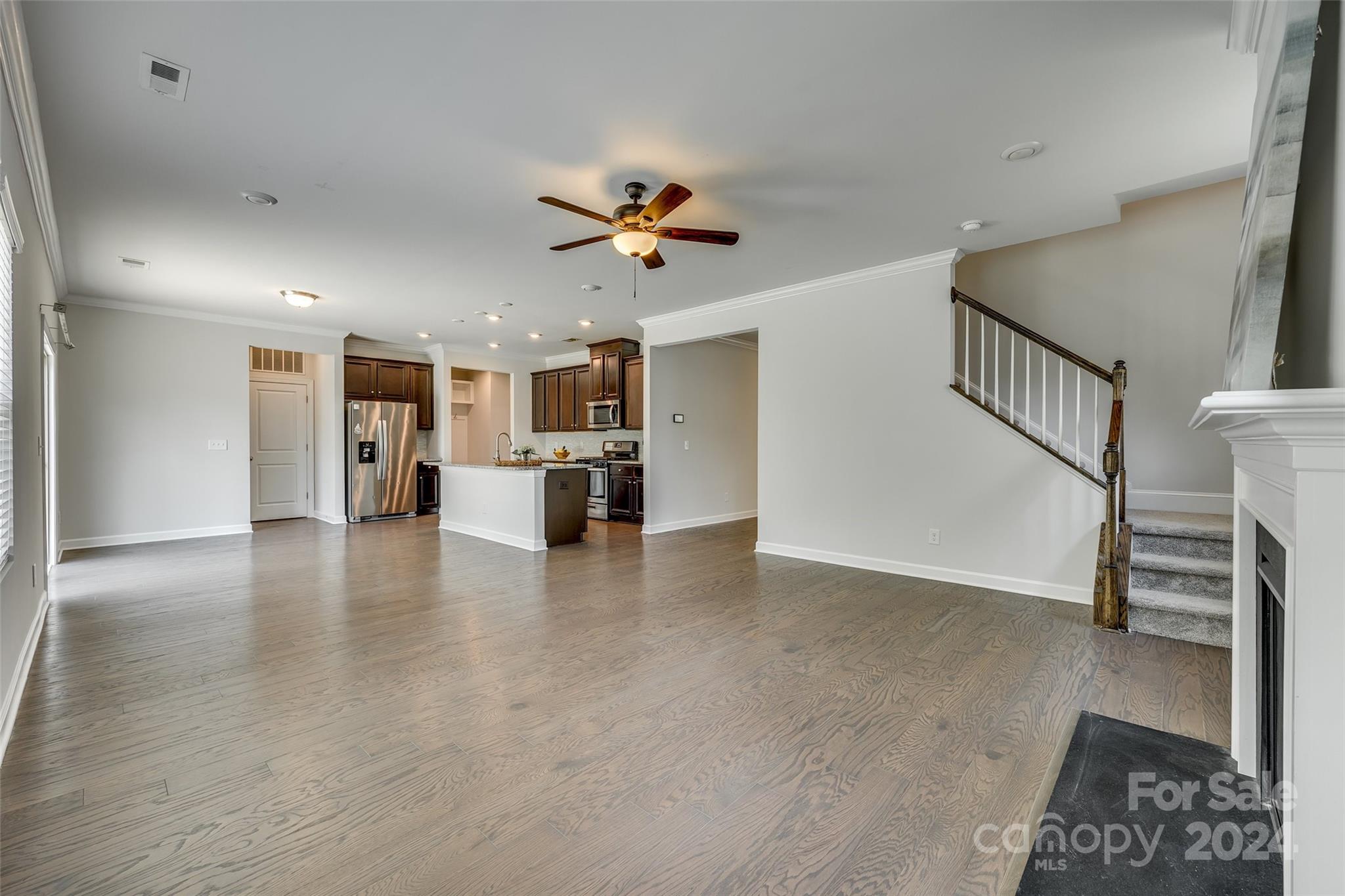 Open living area with light wood floors, a dark-cabinet kitchen, staircase, and fireplace.