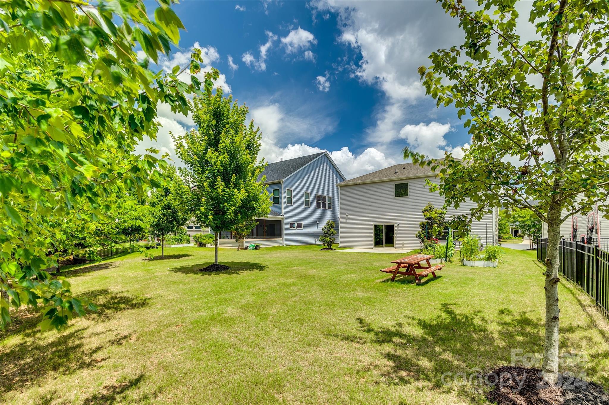 Two-story home behind a leafy backyard with a picnic table and black fencing.
