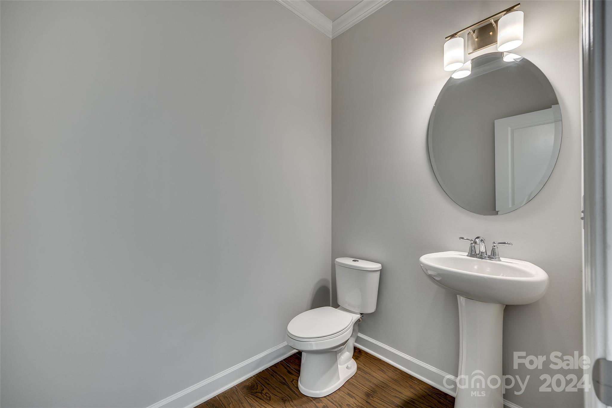 Bathroom with a white pedestal sink, round mirror, toilet, and dark wood flooring.