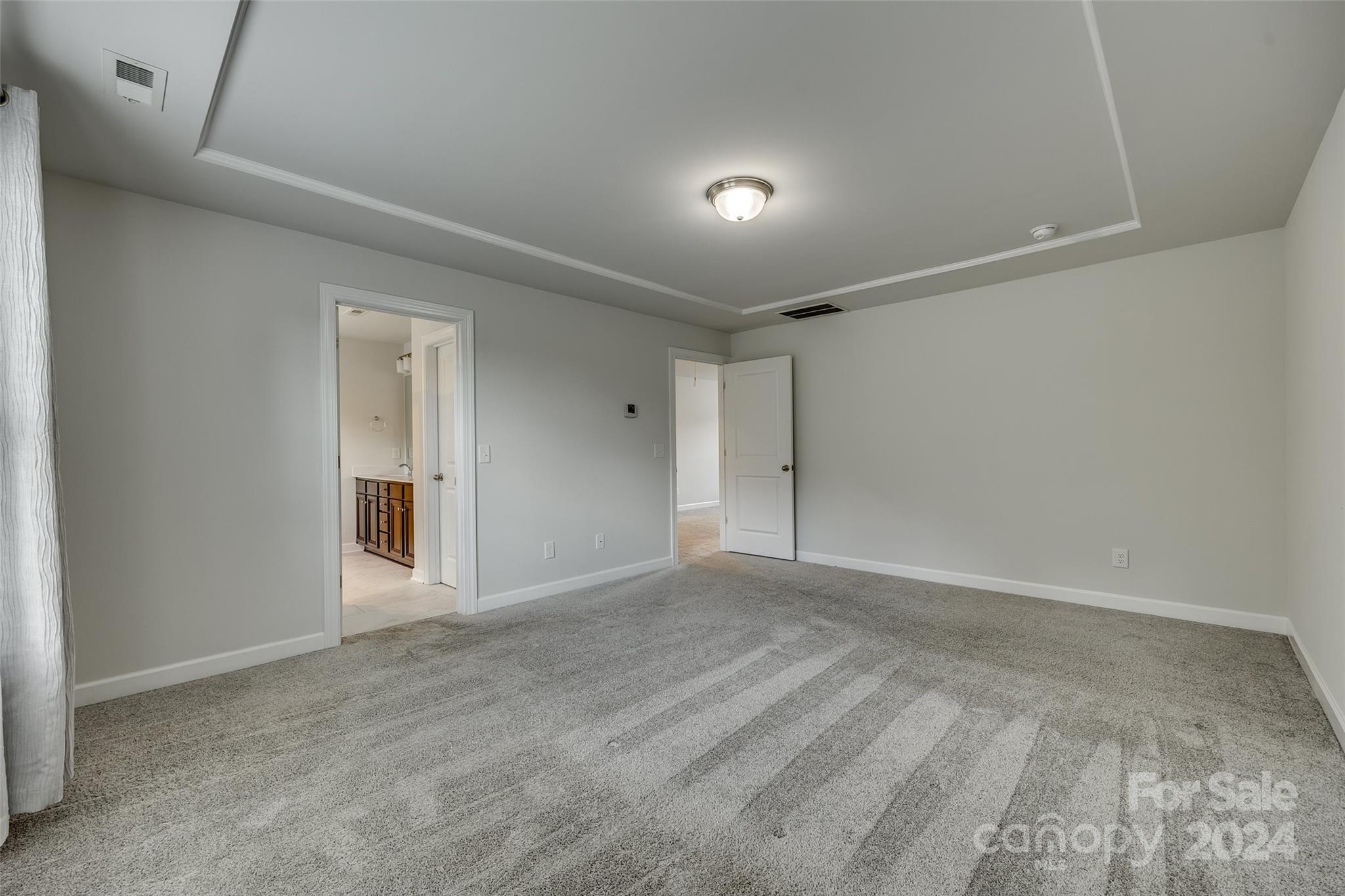 Empty bedroom with gray carpet, tray ceiling, and an open doorway to the bathroom.
