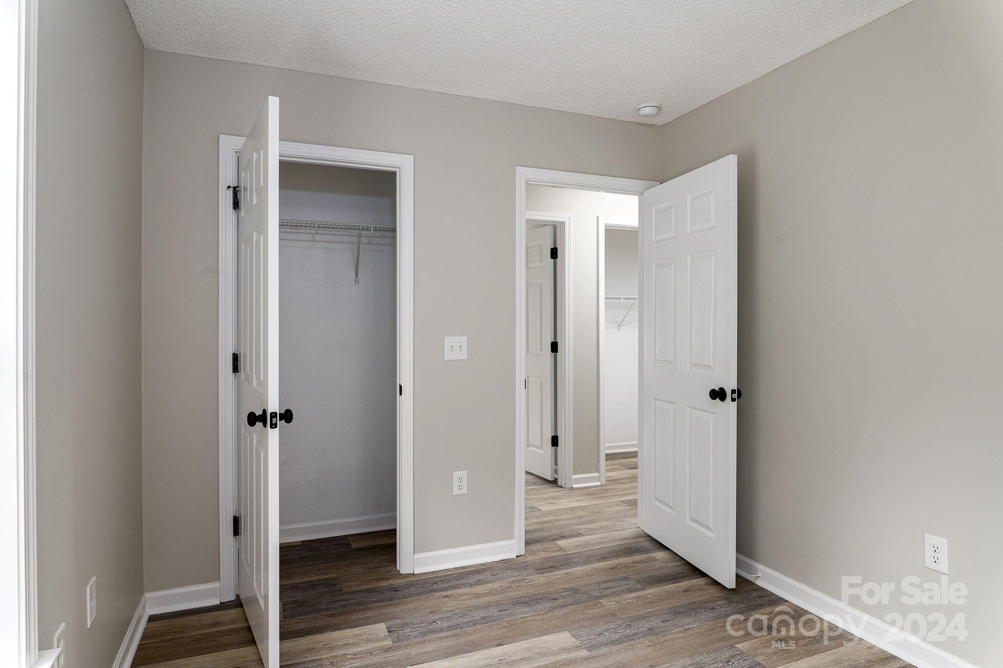 Empty bedroom with wood-look flooring and open doors to closets and a hallway.