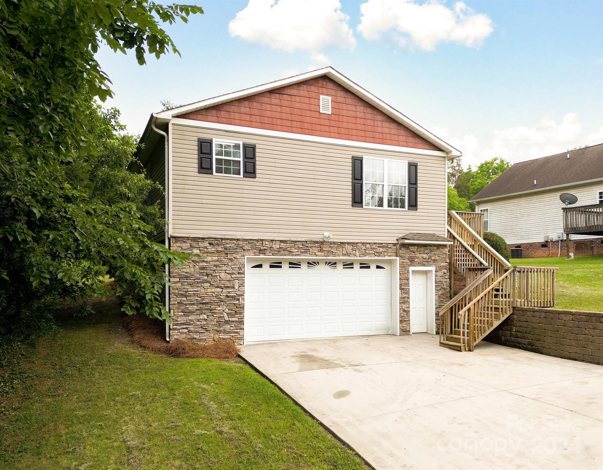 Beige split-level home with a red gable, stone-faced garage, wide driveway, and wooden side stairs.