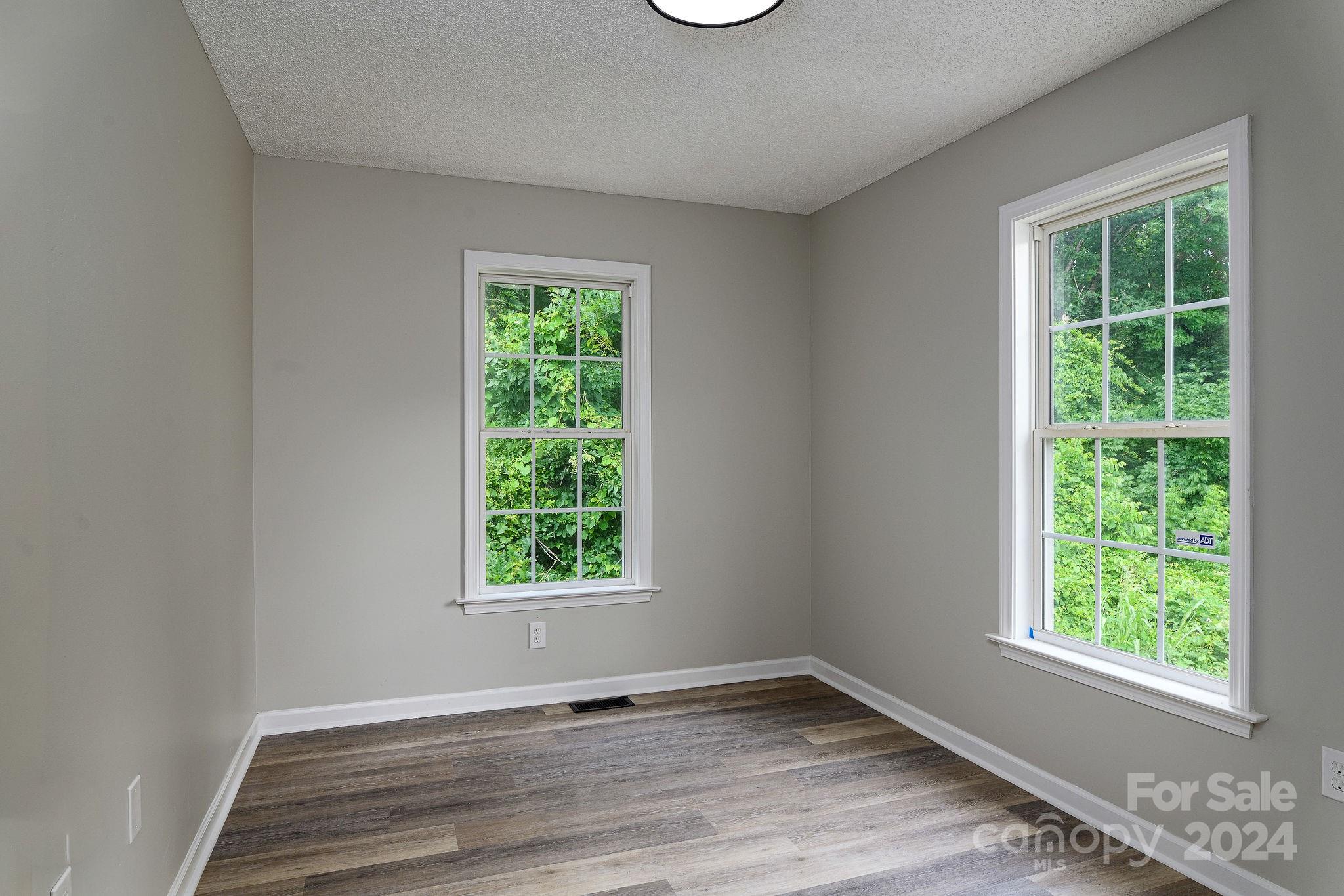 Empty room with gray walls, two leafy-window views, and wood-look plank flooring.