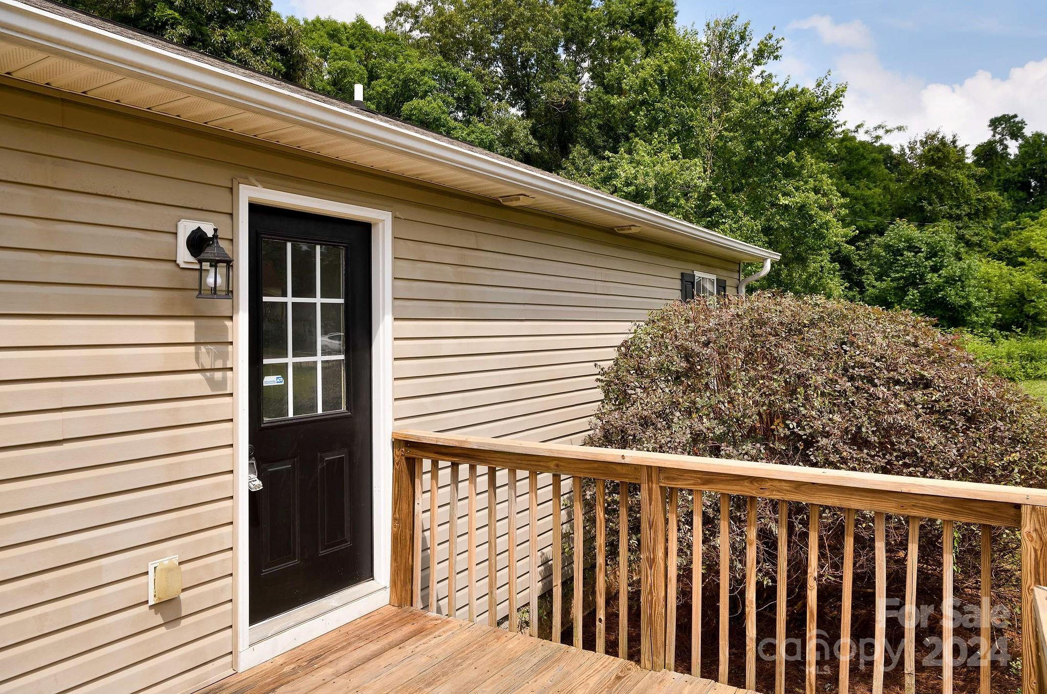 Home’s black side entrance opens onto a wooden deck bordered by shrubs.