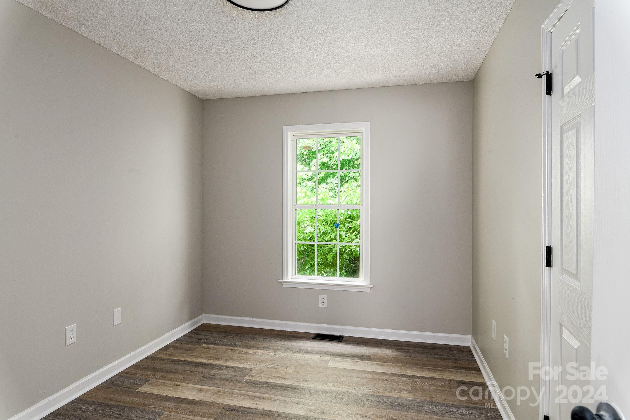Empty room with gray walls, wood-look flooring, and a window facing leafy greenery.