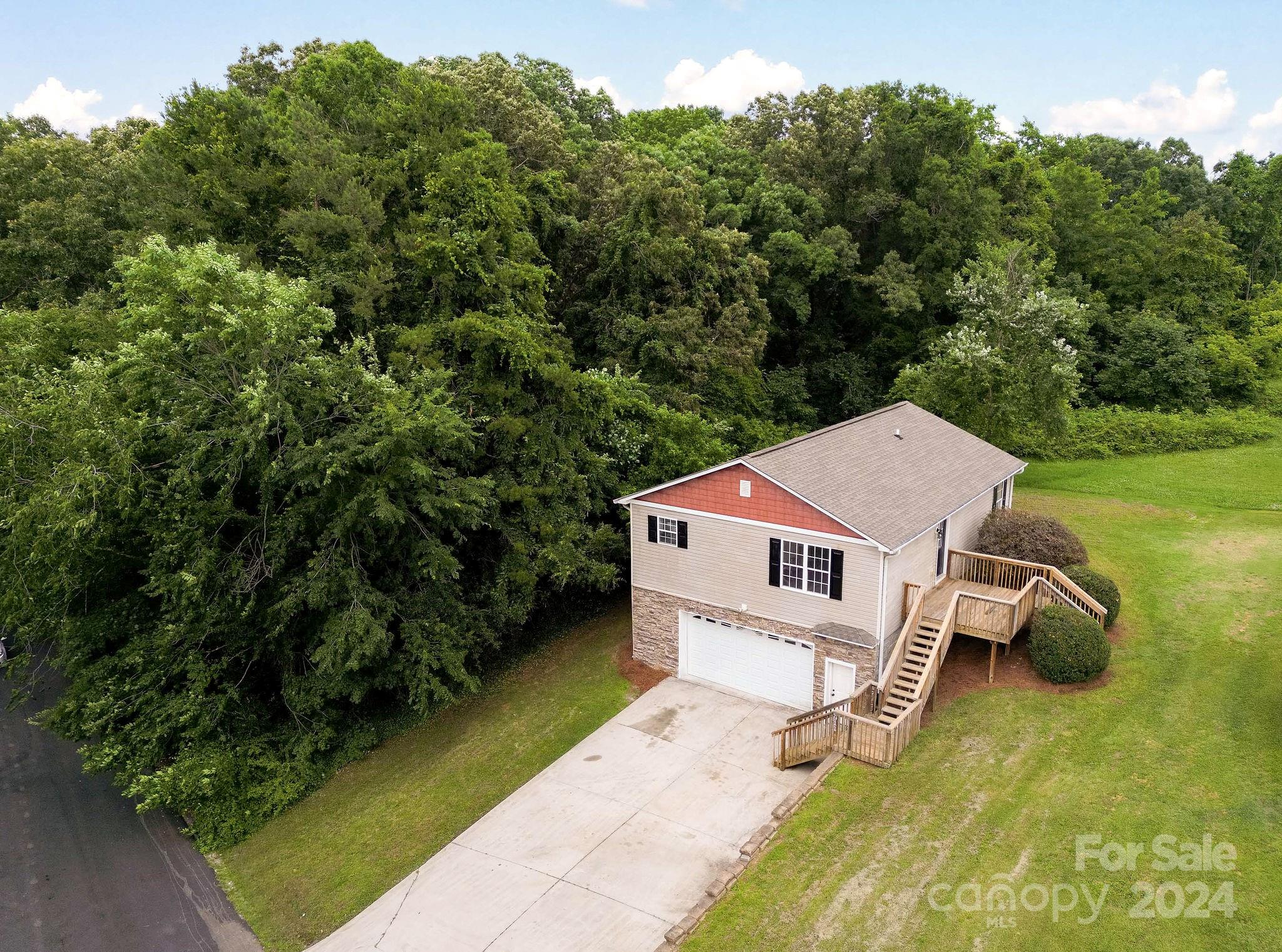 Aerial view of a beige house with a red gable, attached garage, and raised wooden deck beside dense trees.