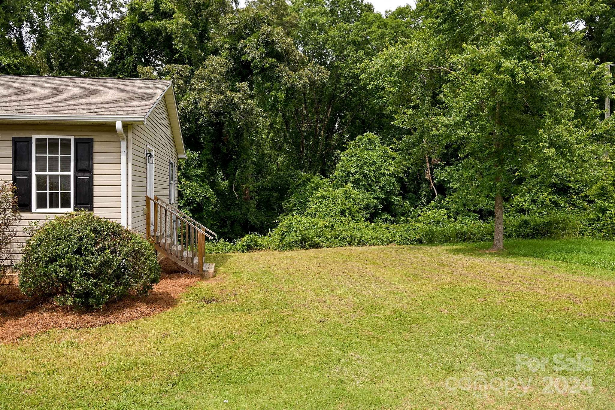 Beige house beside a broad lawn, with a wooden stairway and dense trees behind it.