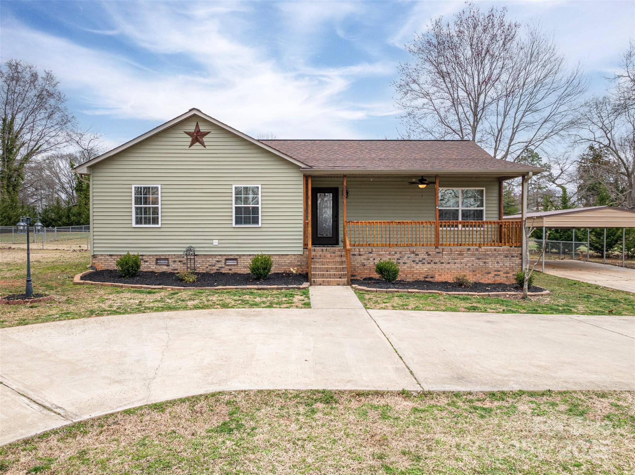 Light-green single-story home with a covered wooden porch, brick foundation, and driveway leading to the front entrance.