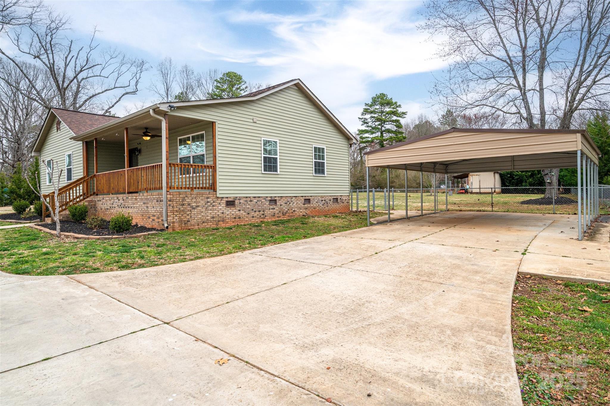 House with a covered wooden porch, brick foundation, and detached metal carport beside a broad driveway.