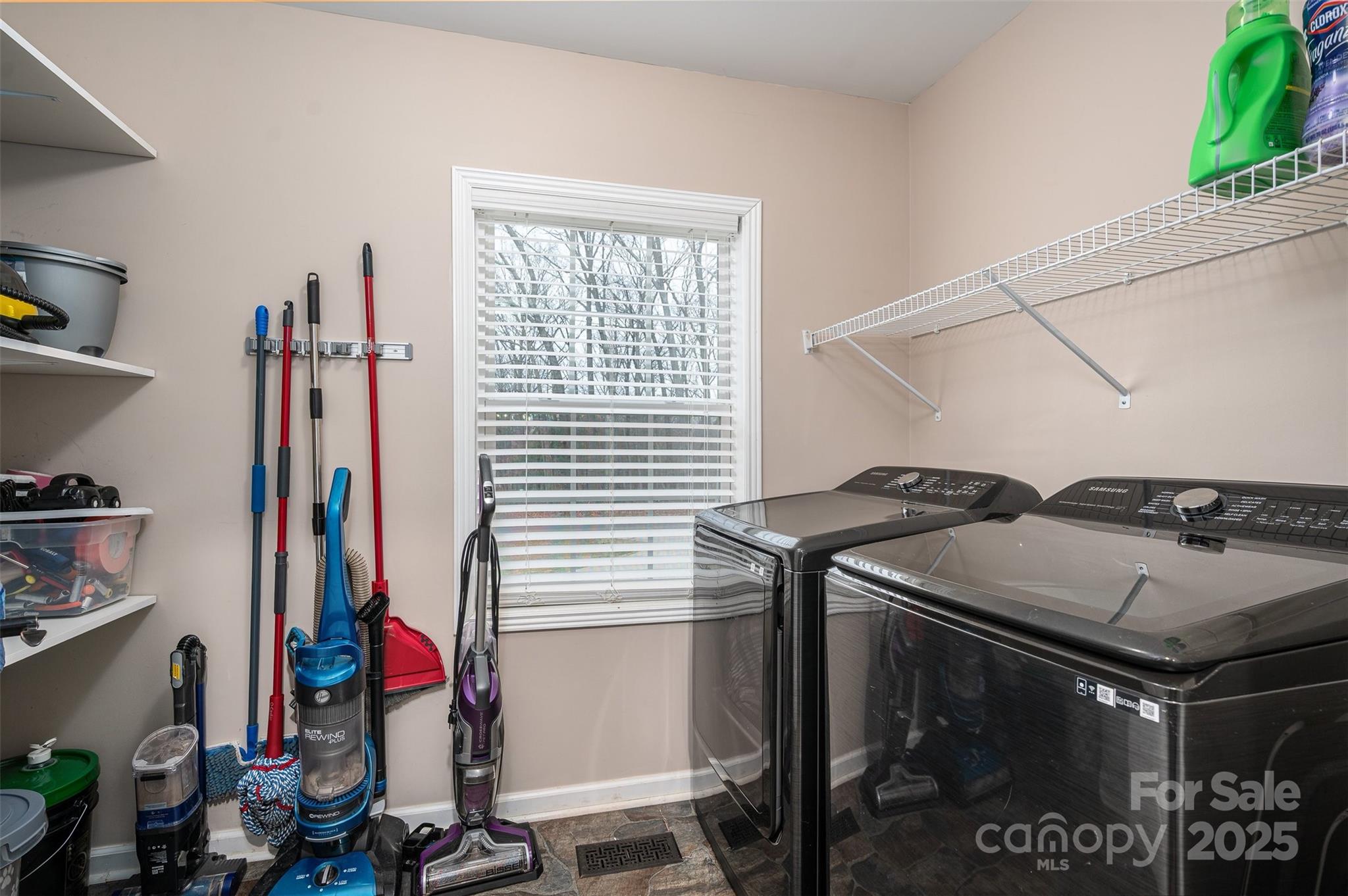 Laundry room with two dark front-loading machines, wire shelving, and cleaning tools beside a window.