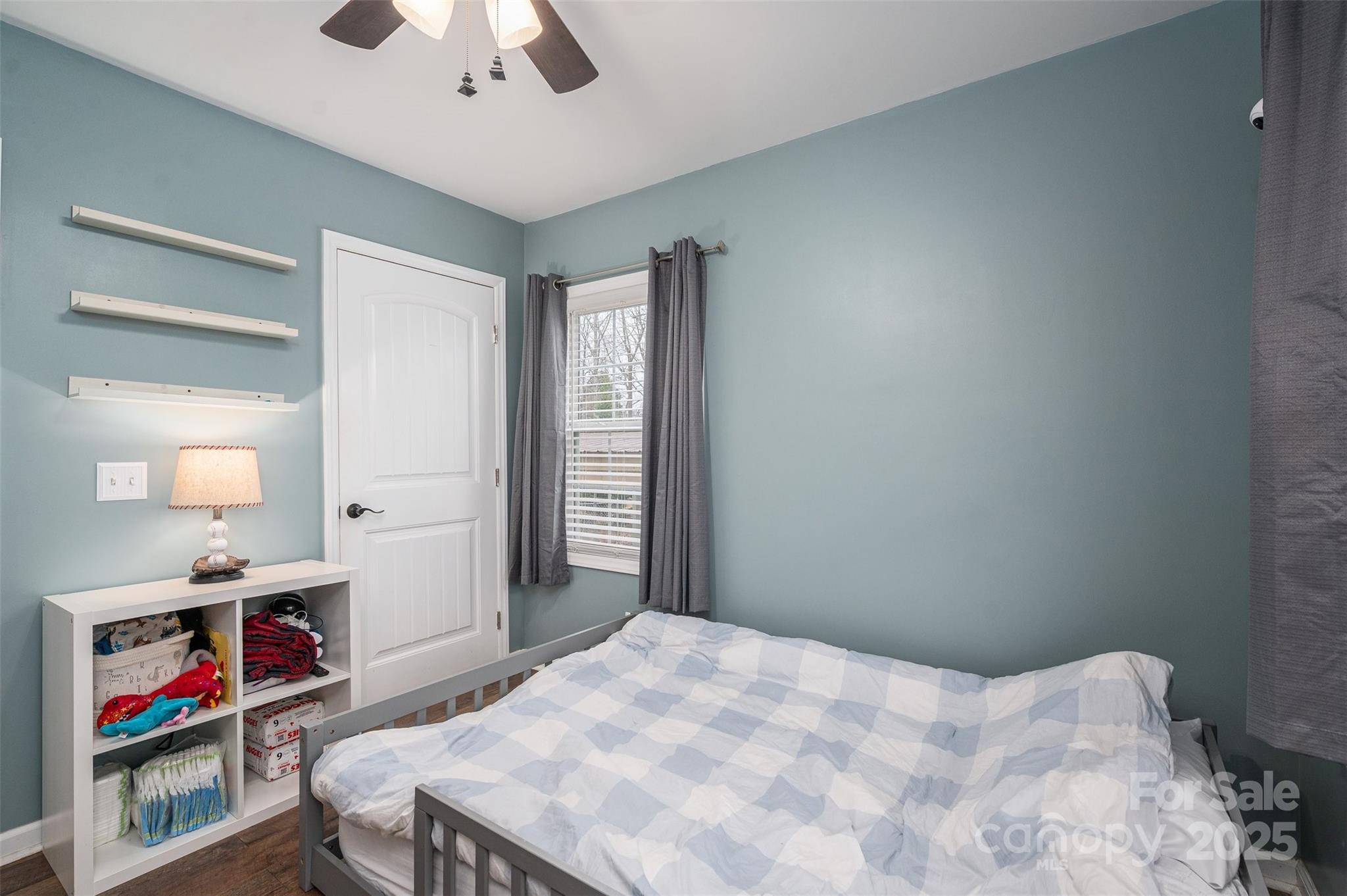 Bedroom with pale blue walls, checkered bedding, gray curtains, and a white cubby shelf.