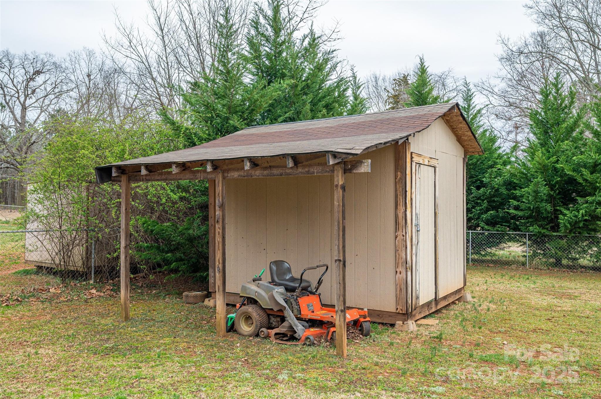 Tan storage shed with a riding mower parked beneath its attached lean-to shelter.