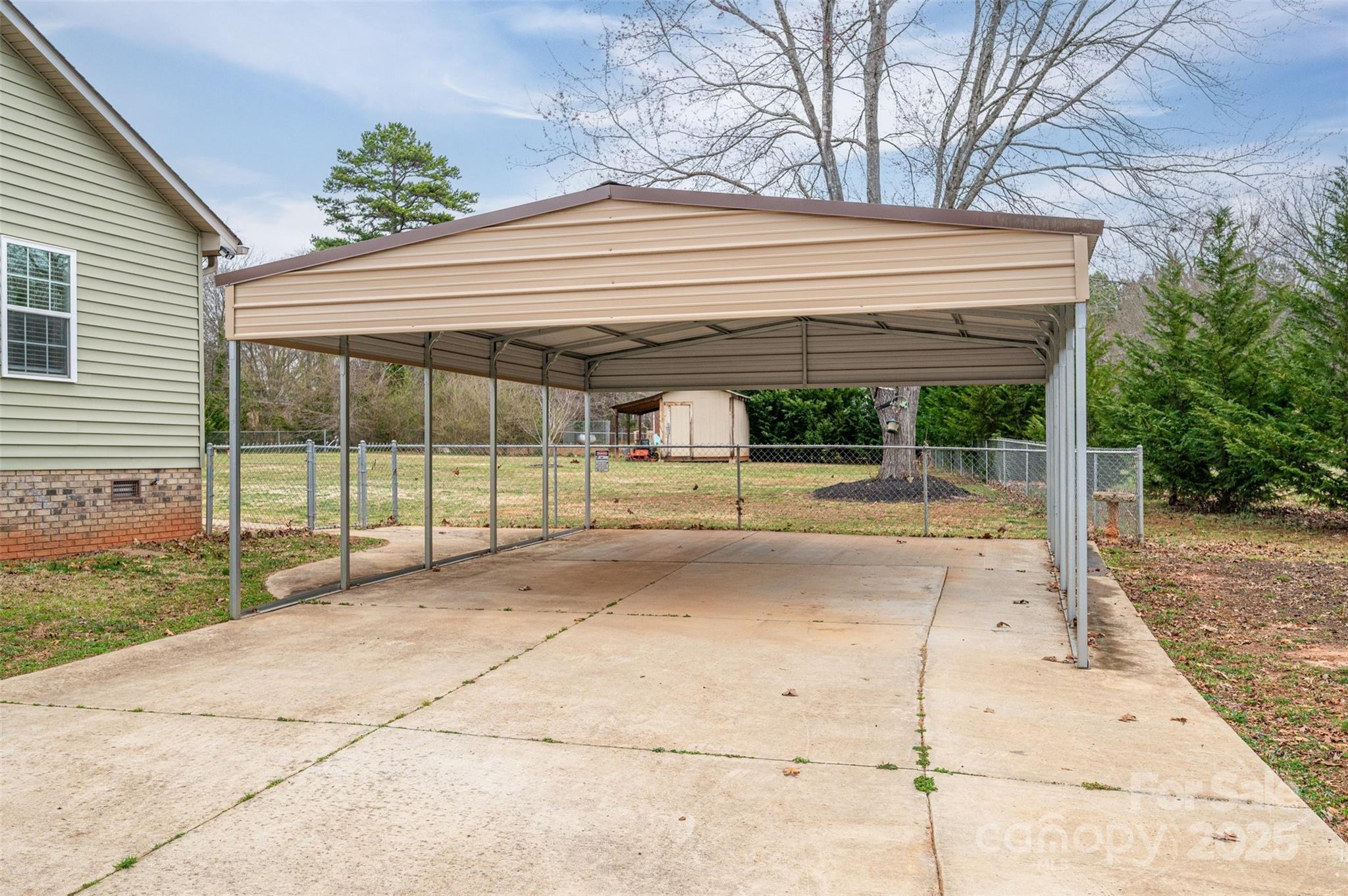 Metal-roofed carport over a broad concrete driveway beside a light green house and fenced yard.