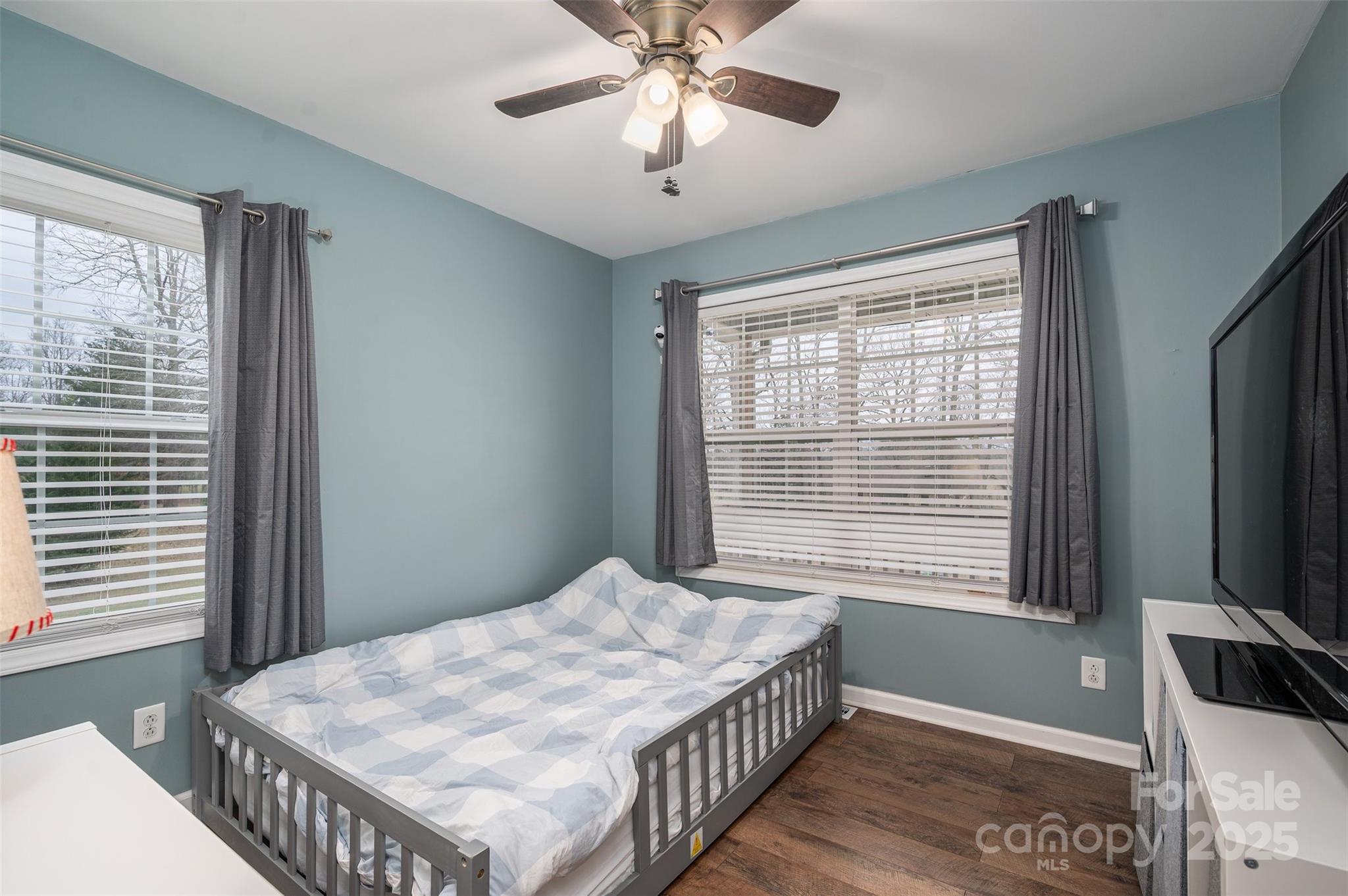 Bedroom with blue-gray walls, a gray bed, checkered bedding, two curtained windows, and dark wood flooring.