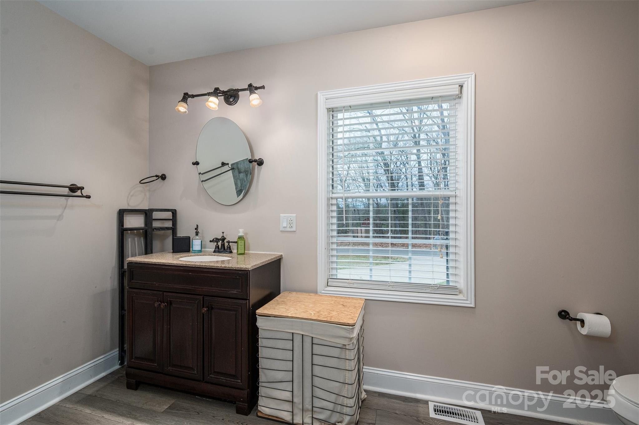 Bathroom with a dark wood vanity, oval mirror, and window overlooking leafless trees.