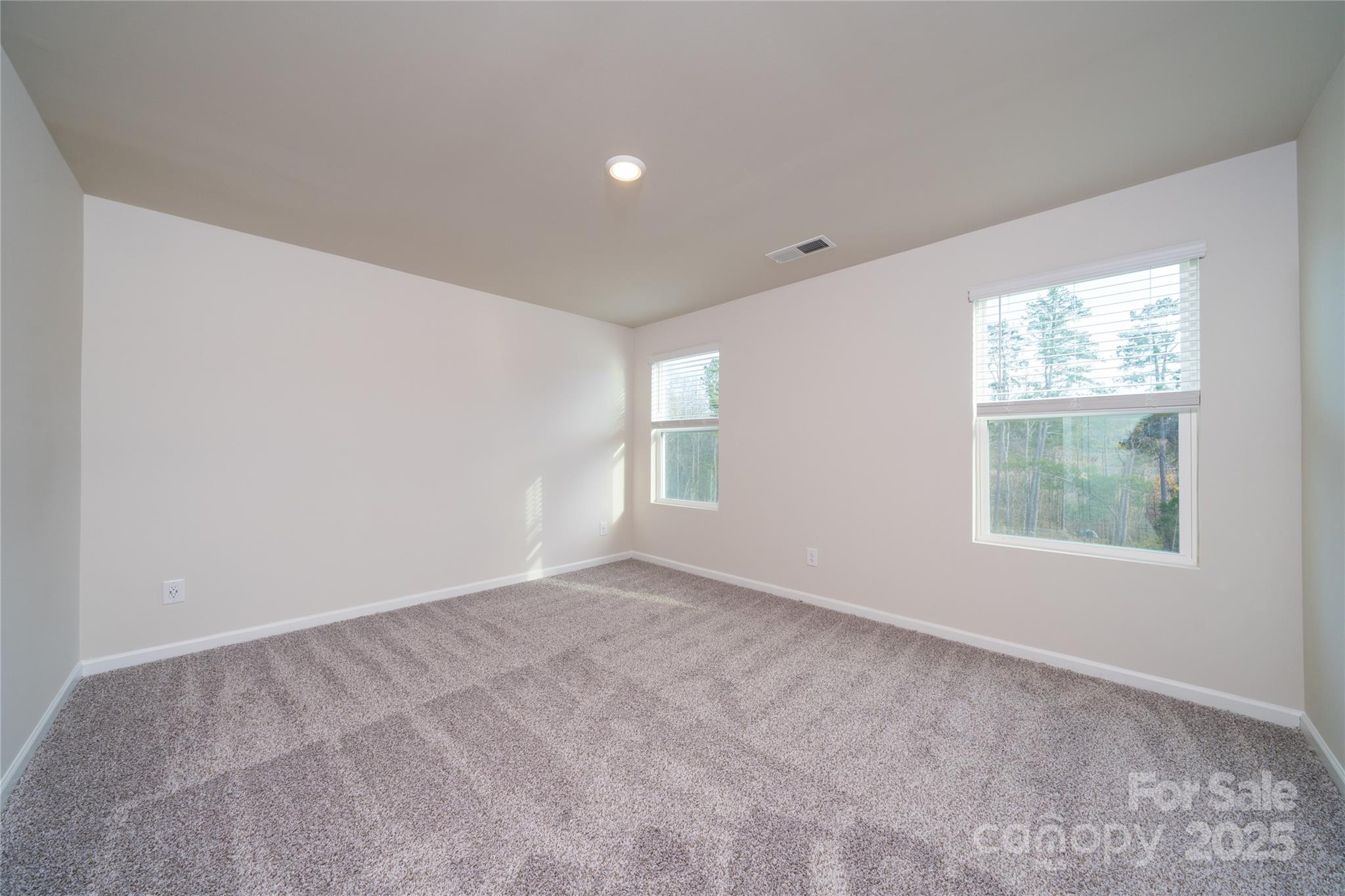 Empty carpeted room with two windows overlooking trees.