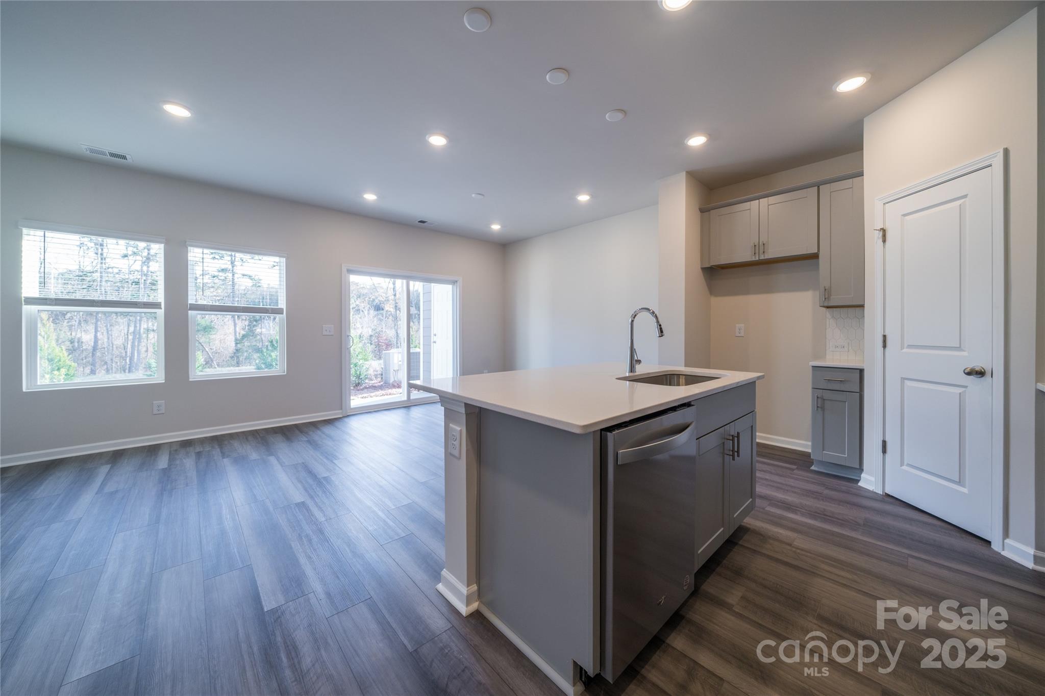 Unfurnished kitchen with a large gray island, sink, dishwasher, and adjoining living area with windows.