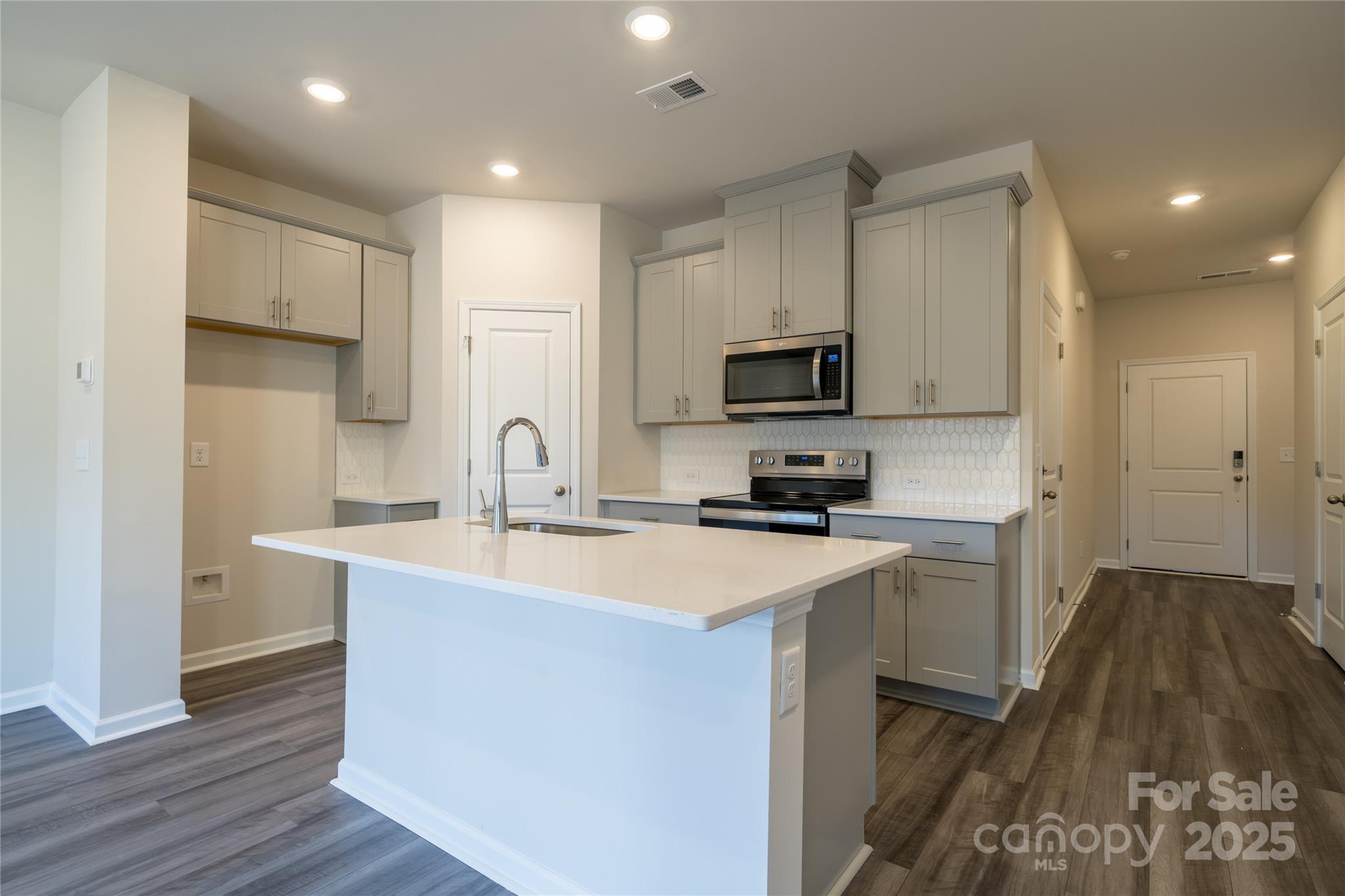 Kitchen with gray cabinets, a large white island, stainless appliances, and a hallway to the right.