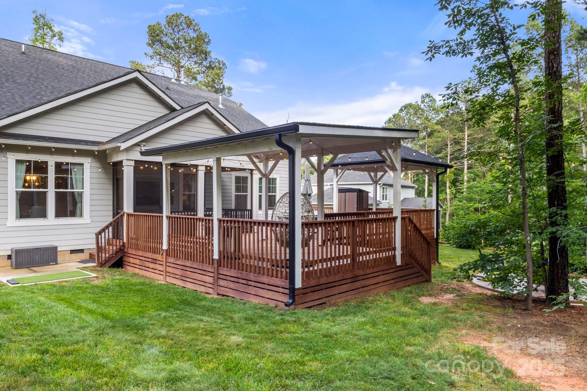 Home’s raised wooden deck with a covered pergola and string lights, bordered by a wooded yard.