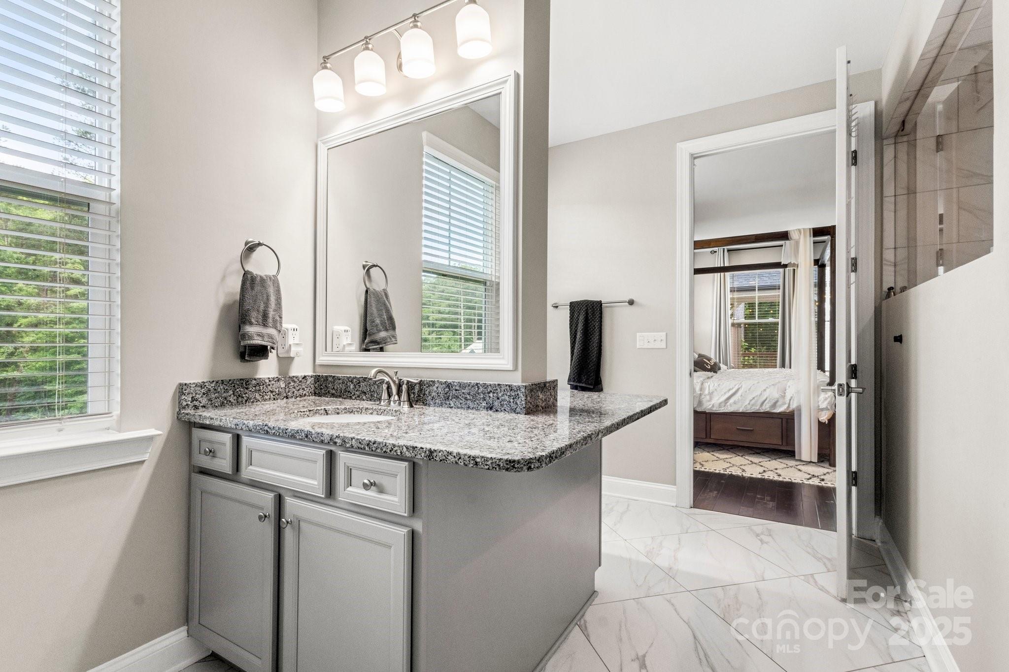 Bright bathroom with a granite-topped gray vanity, glass shower, and doorway to a bedroom.