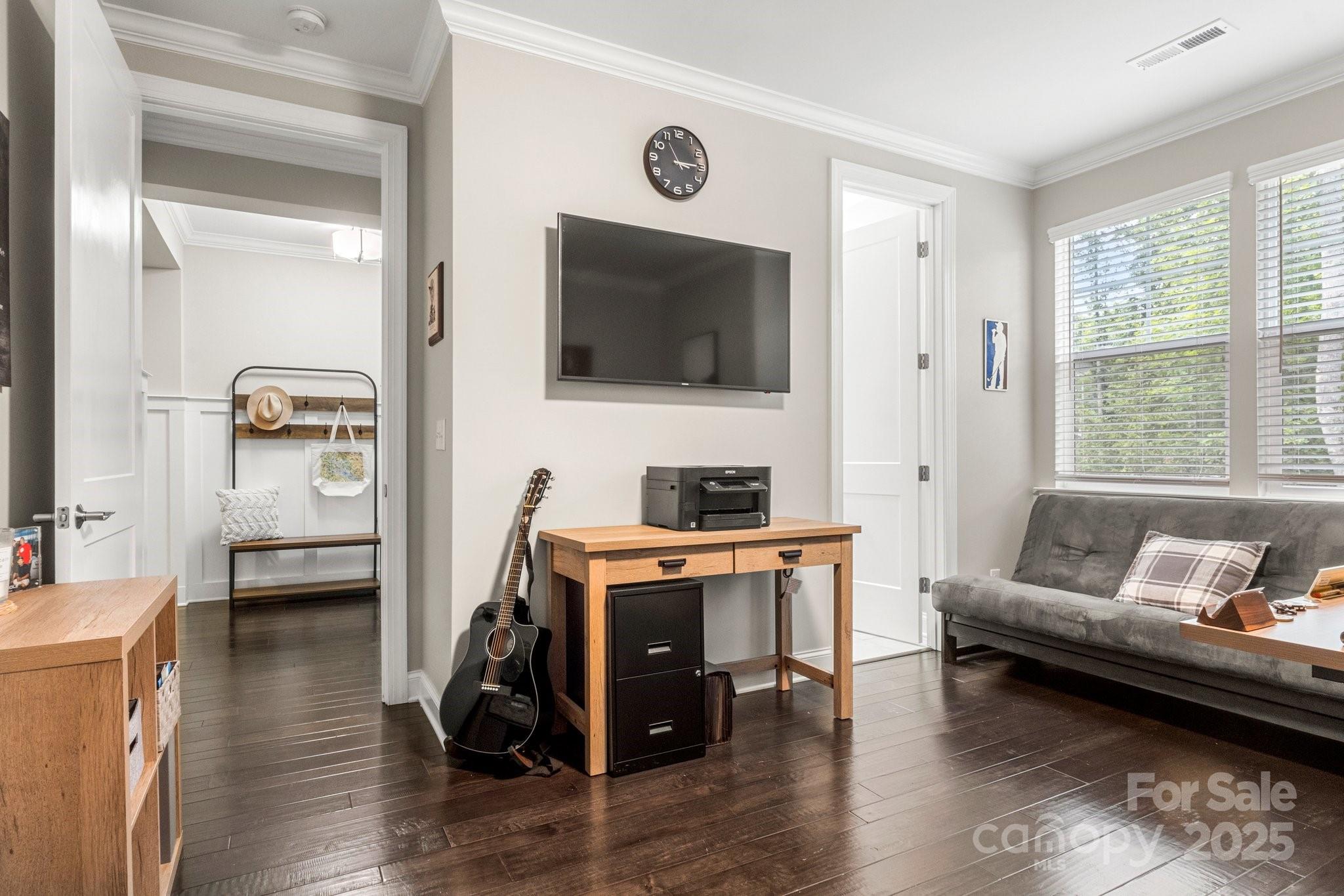 Furnished home office with a wooden desk, black guitar, wall-mounted TV, and gray sofa by the windows.