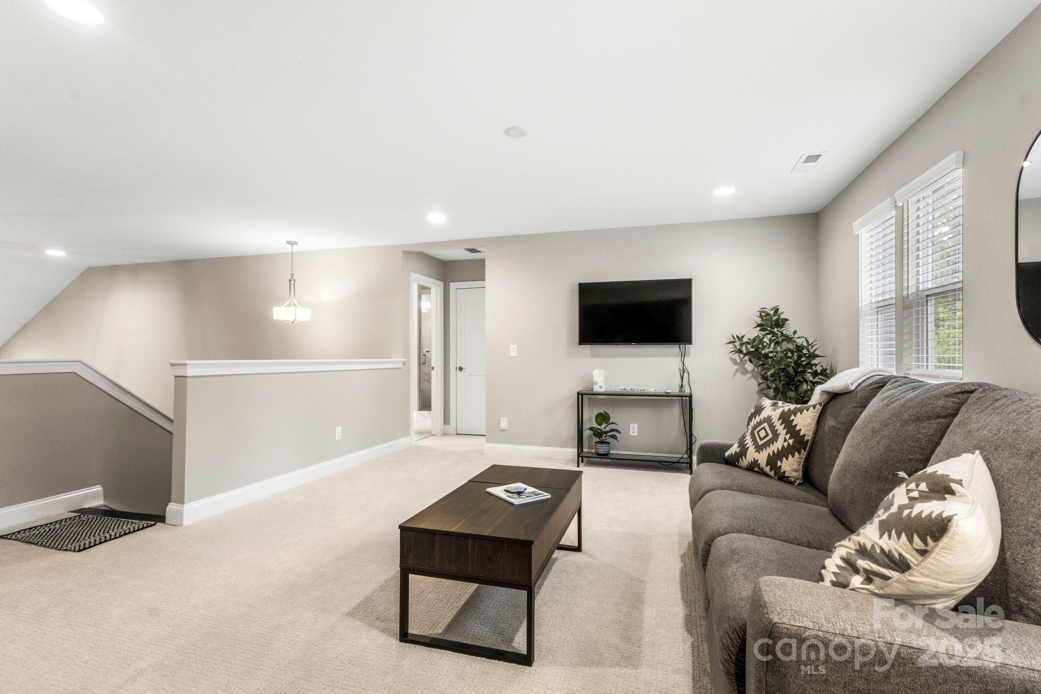 Staged upstairs living room with a gray sectional, wall-mounted TV, and stairway.
