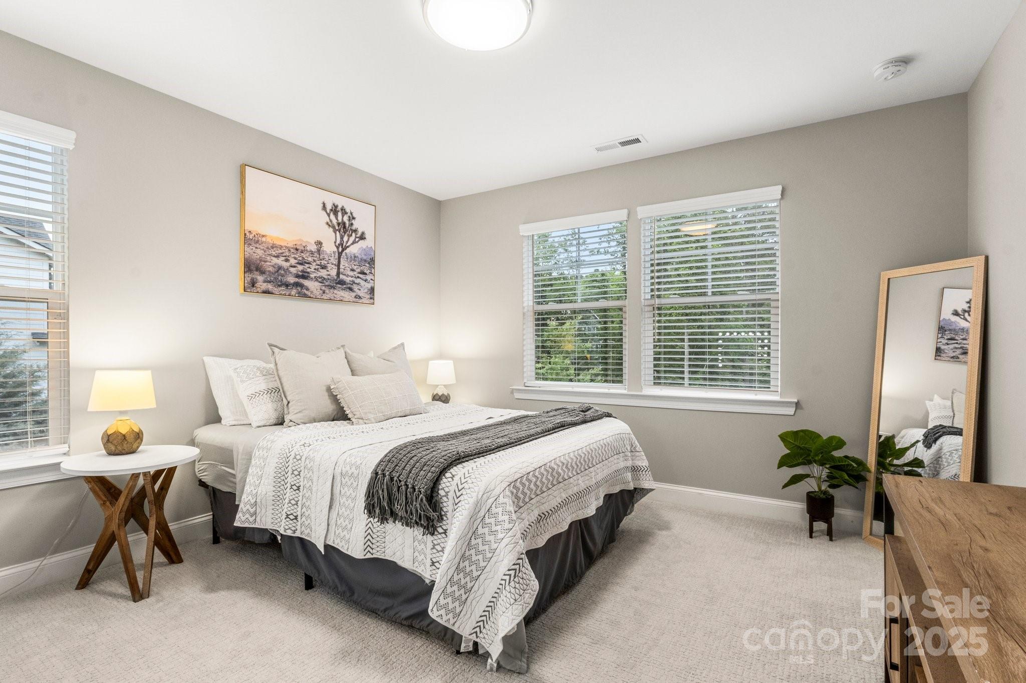 Staged bedroom with patterned white bedding, a gray throw, and windows overlooking greenery.