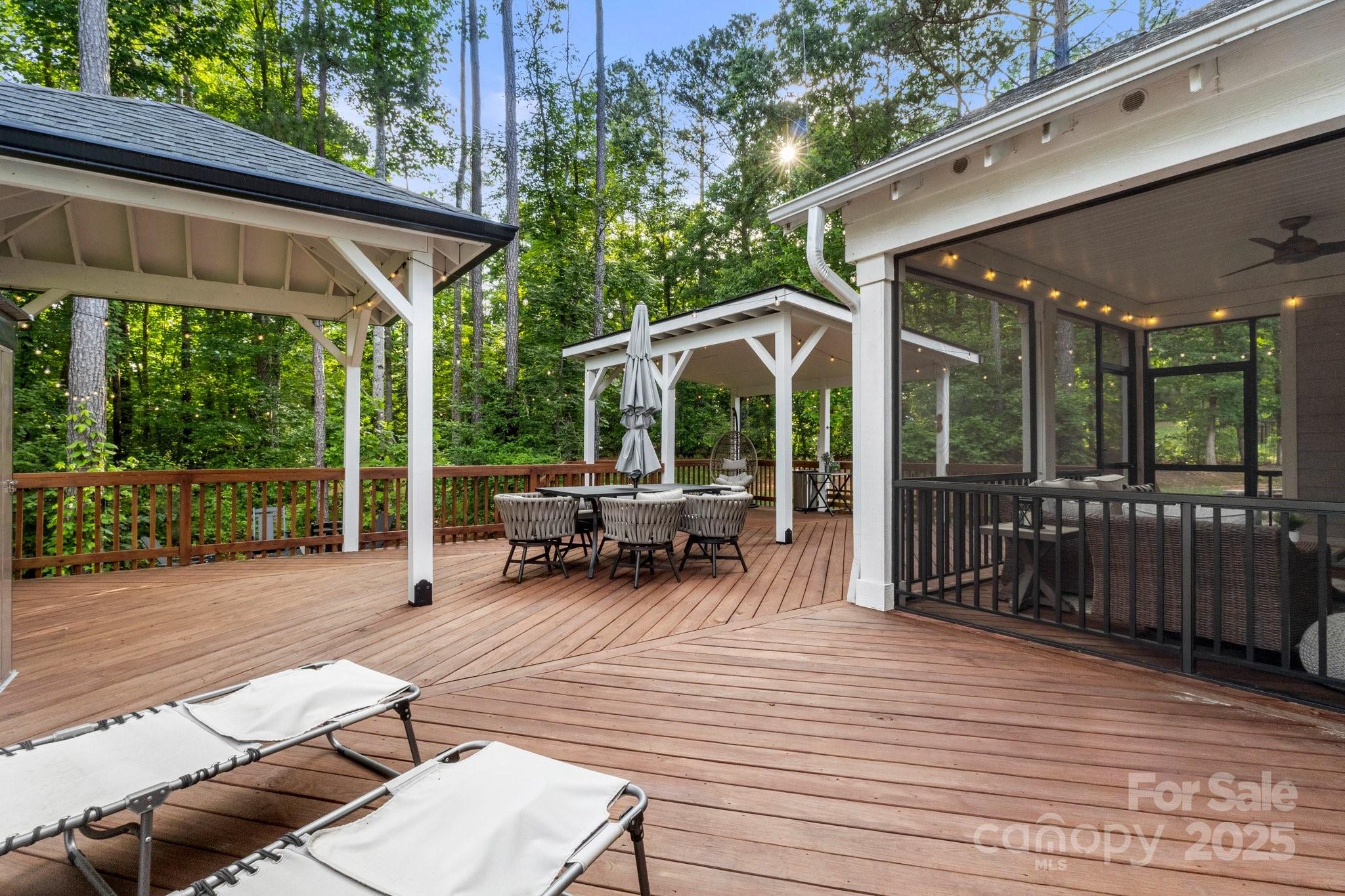 Wood deck with covered pergolas, outdoor dining set, lounge chairs, and screened porch among trees.