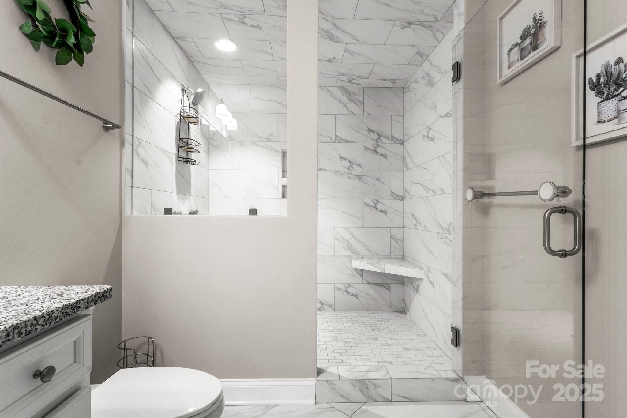 Bathroom with a glass-enclosed marble-look tile shower and built-in bench beside the toilet and vanity.