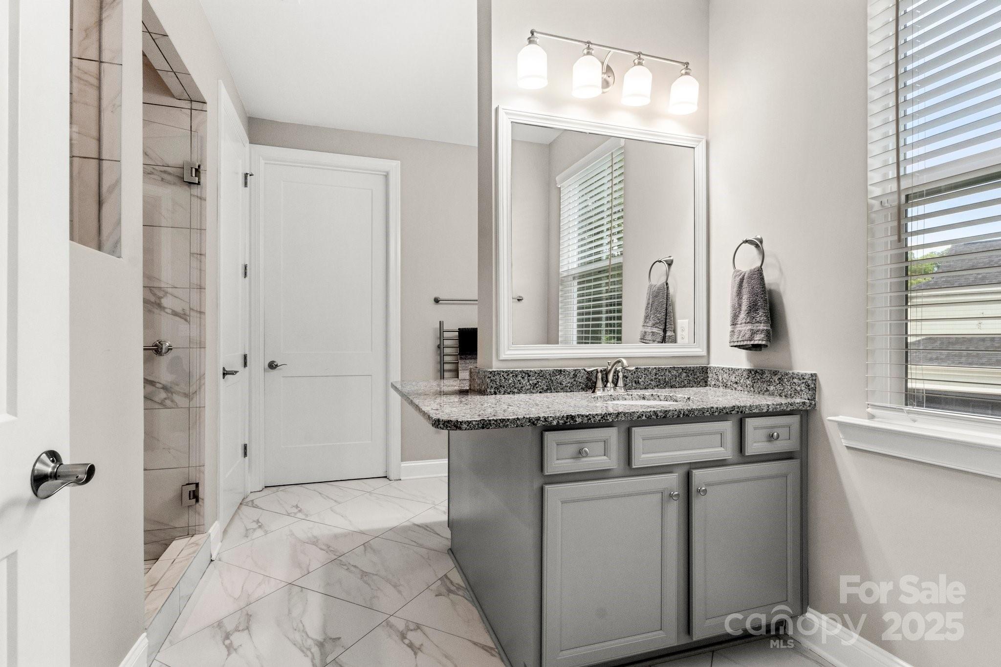 Bathroom with a gray vanity and granite counter beside a marble-look tiled shower and floor.